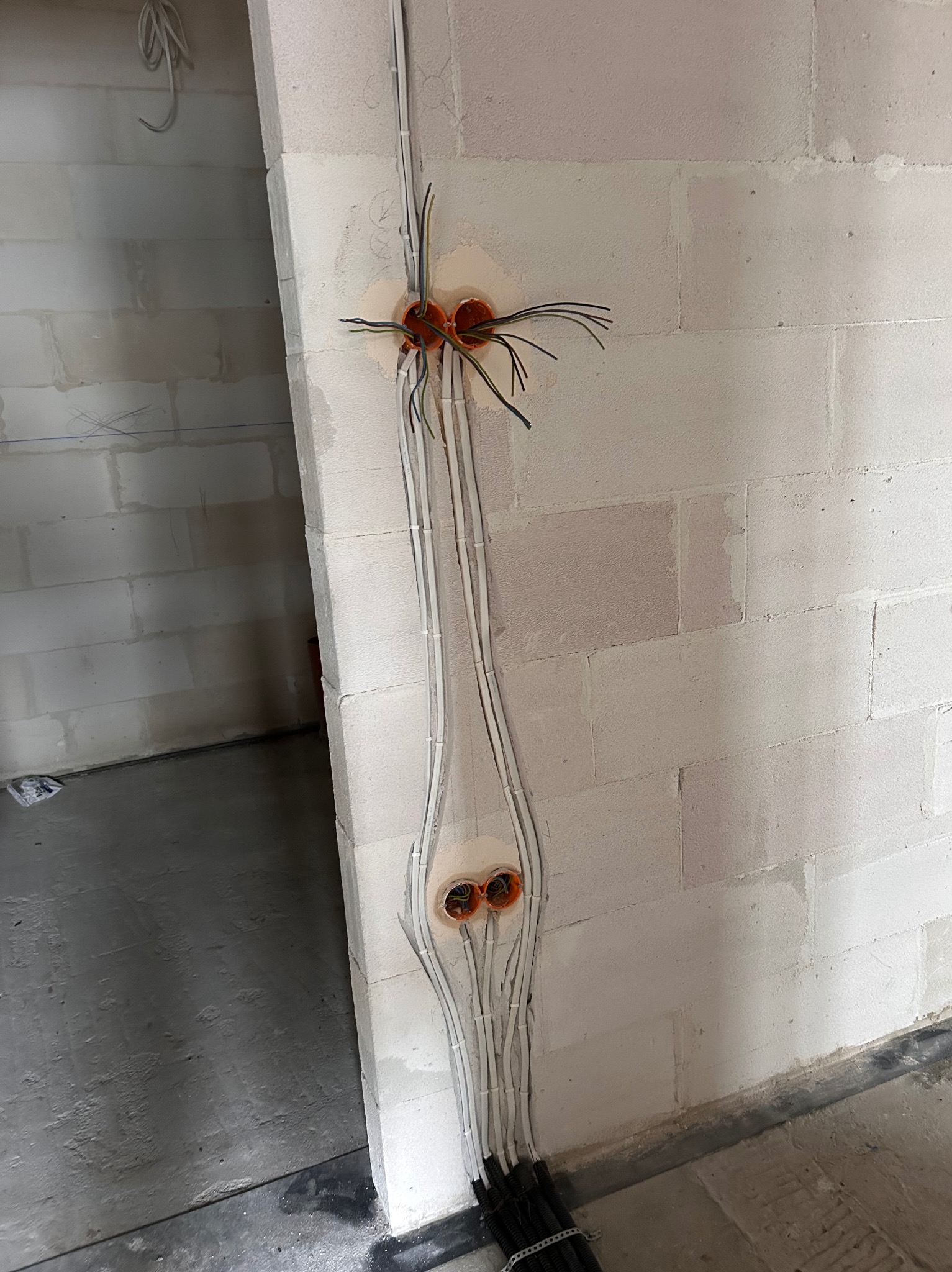 Instalacja elektryczna w surowym wnętrzu: białe przewody prowadzone po ścianie z bloczków, pomarańczowe puszki podtynkowe. Widok na wczesny etap prac.