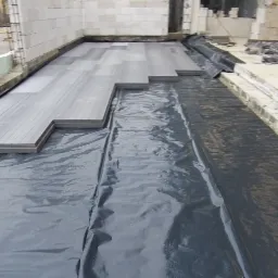 Układanie szarej, kompozytowej podłogi tarasowej na czarnej folii hydroizolacyjnej w surowym stanie, widoczne bloczki betonowe ścian i przygotowane podłoże.