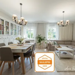 Jasne, przestronne wnętrze salonu z jadalnią: stół z szarymi krzesłami, duży narożnik, biała biblioteczka i wiszące lampy w stylu industrialnym.