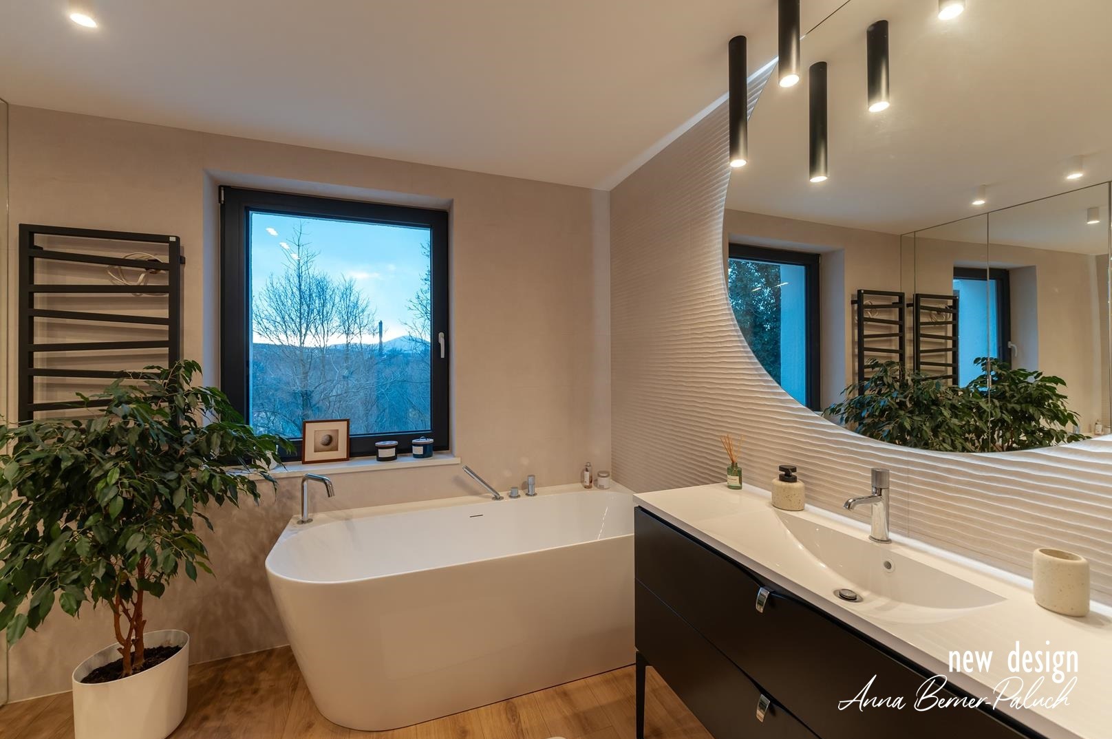 Nowoczesna łazienka z wolnostojącą wanną przy oknie z widokiem na góry, umywalką nablatową na czarnej szafce, okrągłym lustrem z dekoracyjną ścianą i czarnym grzejnikiem drabinkowym.