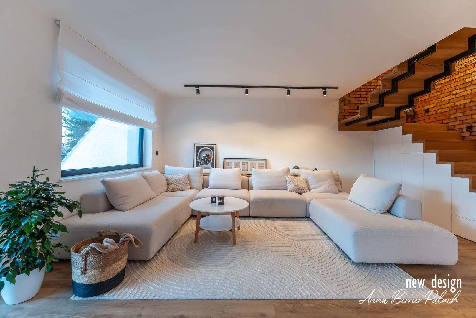 Jasne wnętrze salonu z modułową sofą, okrągłym stolikiem kawowym i schodami z ceglanym akcentem, oświetlone reflektorami szynowymi.