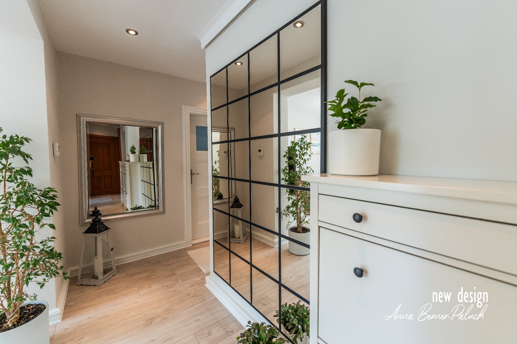 Nowoczesny przedpokój z dużym lustrem w czarnej ramie, białą komodą i roślinami doniczkowymi; widoczne odbicie wnętrza w lustrze.