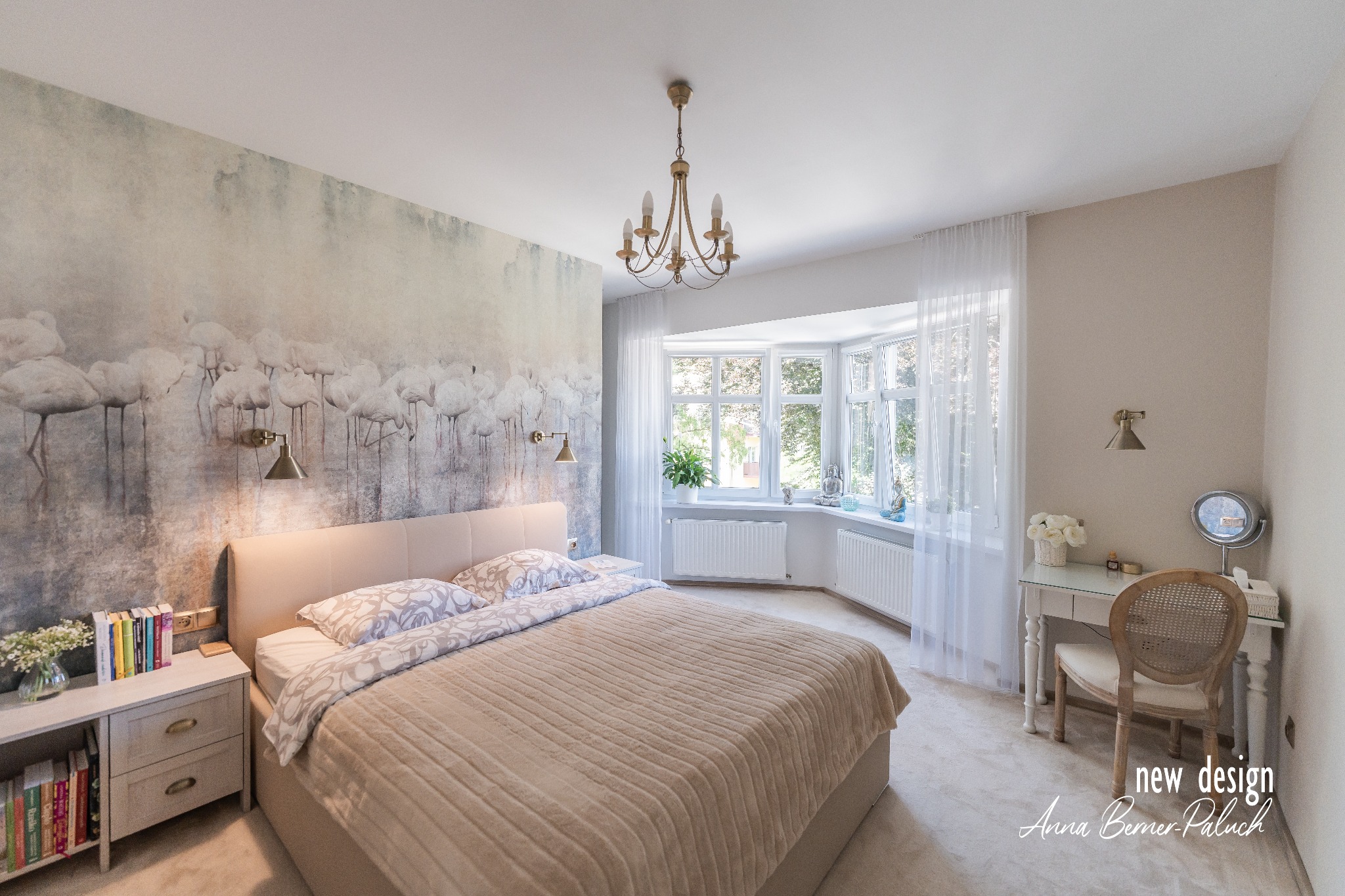 Jasna sypialnia z łóżkiem, toaletką i ścianą z motywem flamingów, oświetlona złotym żyrandolem i lampami ściennymi.