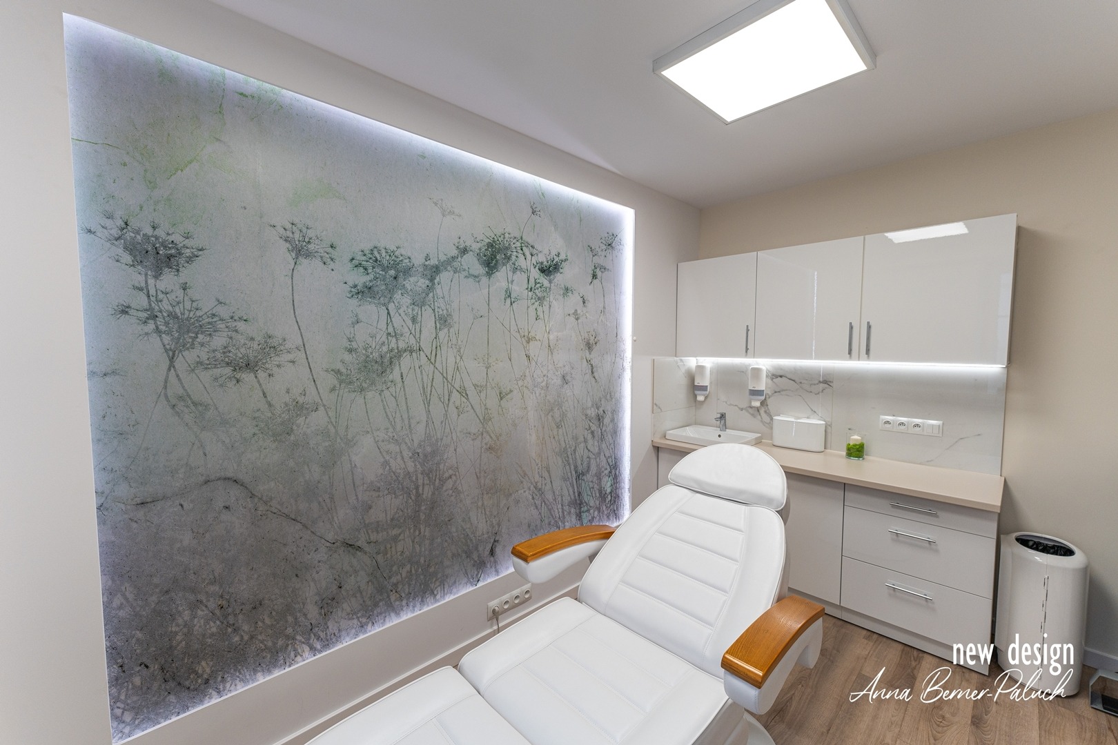 Biały fotel kosmetyczny z drewnianymi podłokietnikami w jasnym gabinecie z umywalką, białymi szafkami i ścianą z motywem roślinnym podświetloną ledami.