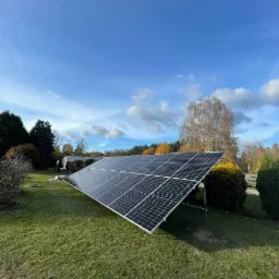 Panel fotowoltaiczny zamontowany na trawniku przed domem w słoneczny dzień z błękitnym niebem.