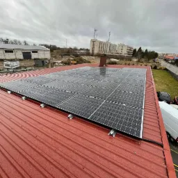Panele fotowoltaiczne na czerwonym, metalowym dachu budynku przemysłowego w pochmurny dzień, widok z góry.