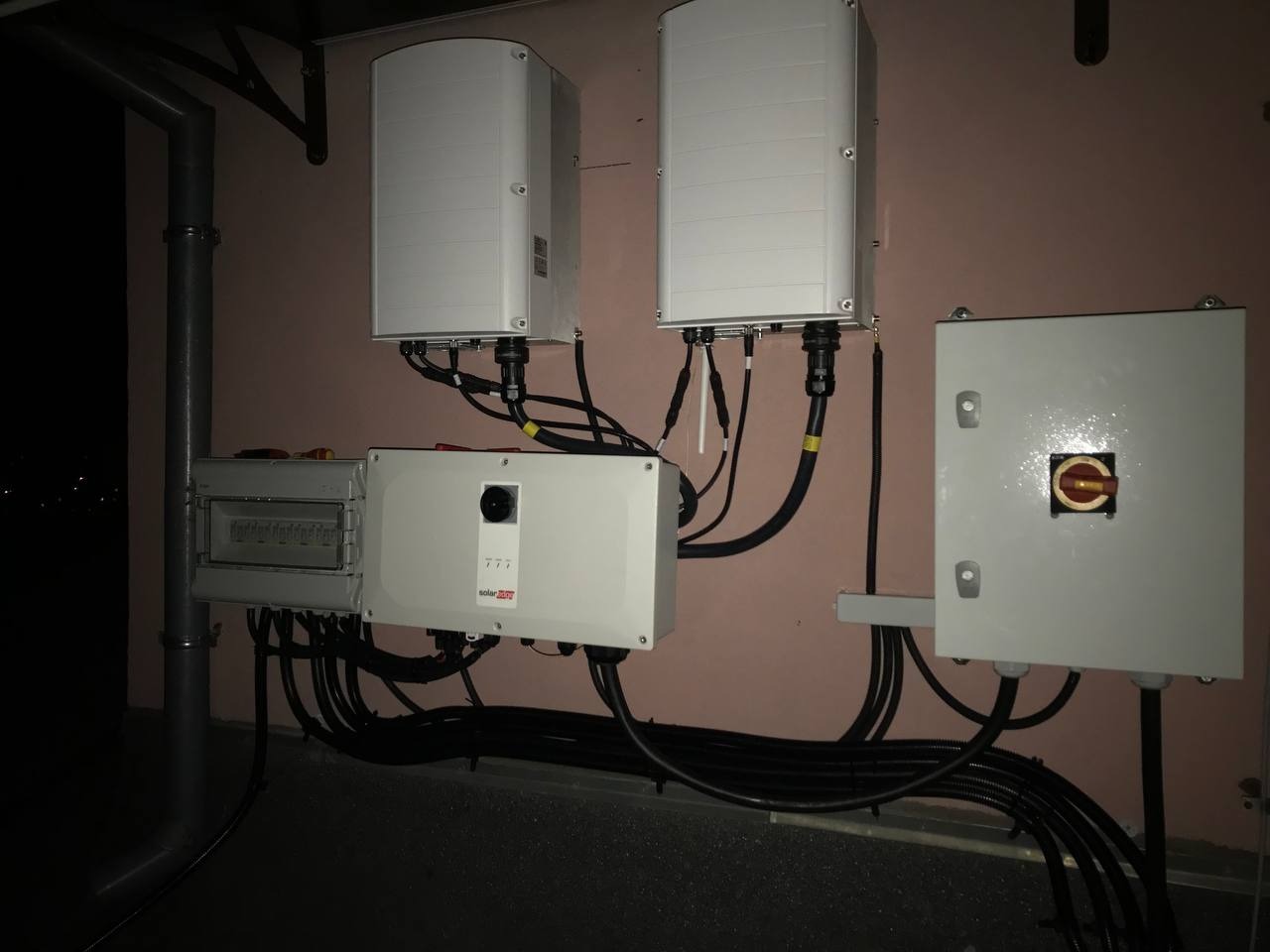 Zewnętrzna skrzynka rozdzielcza, falownik SolarEdge i dwa inwertery na ścianie budynku z widocznymi kablami w peszlach.