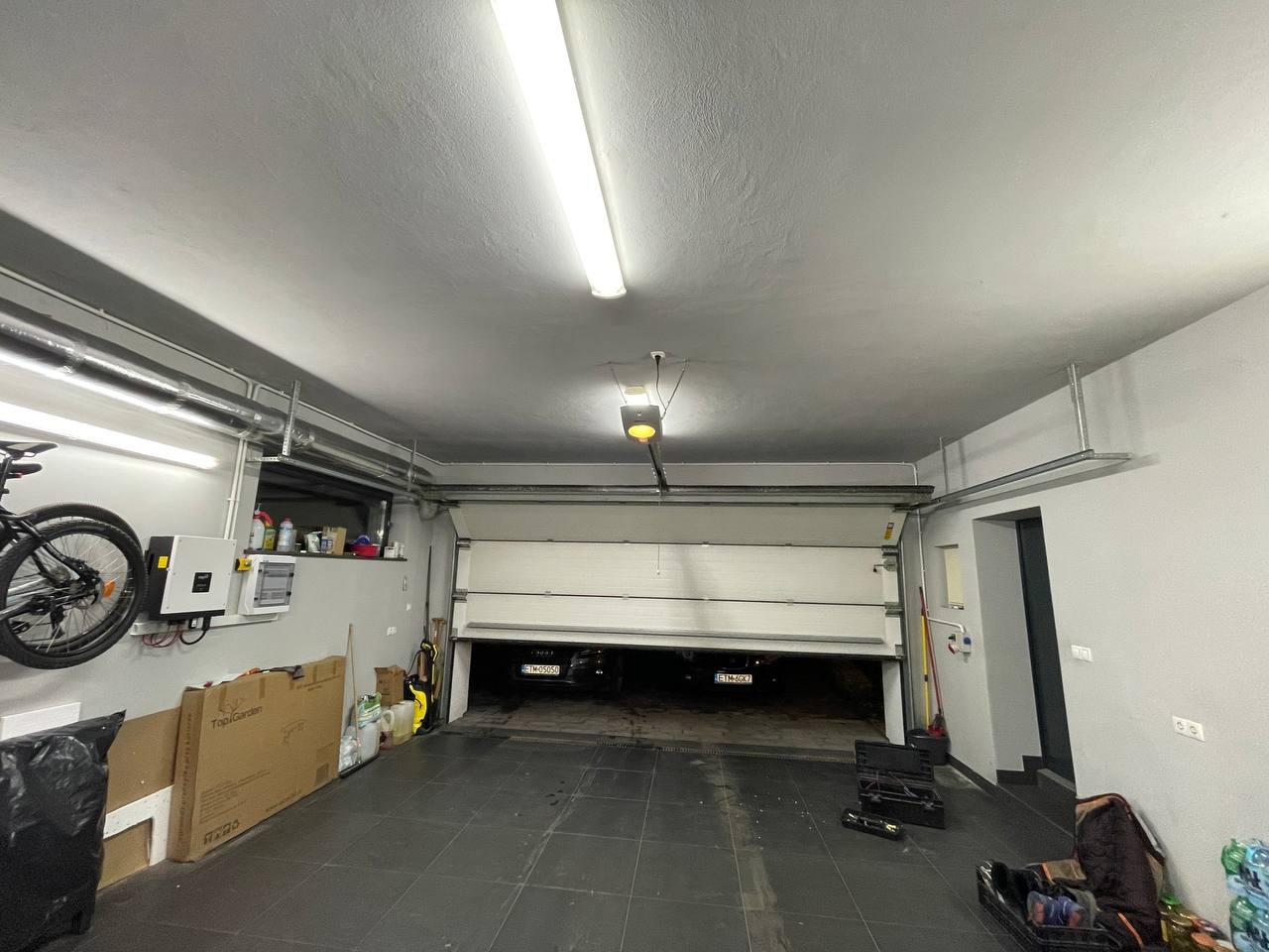 Częściowo otwarta segmentowa brama garażowa z widocznym mechanizmem automatycznego otwierania, instalacją elektryczną na ścianie, rowerem i narzędziami w garażu z szarą podłogą i ścianami.
