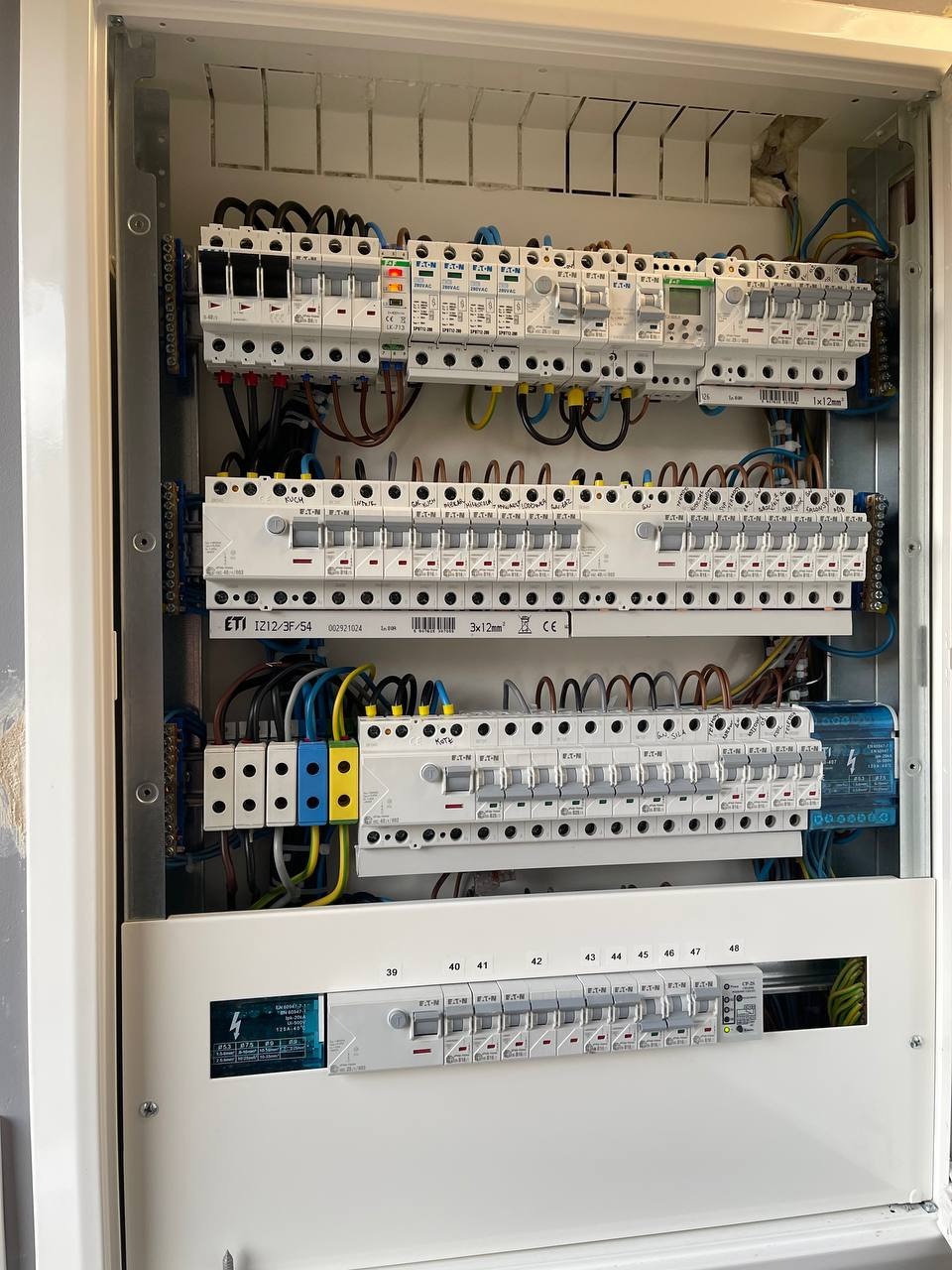 Otwarta skrzynka rozdzielcza z bezpiecznikami i okablowaniem, widoczne różne typy zabezpieczeń i oznaczenia przewodów, w tym żółto-niebieskie.