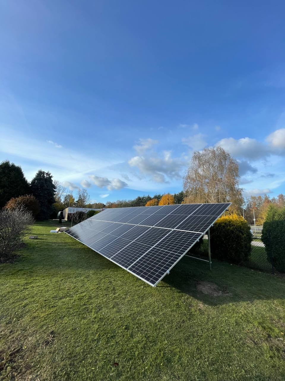 Panel fotowoltaiczny zamontowany na trawniku przed domem w słoneczny dzień z błękitnym niebem.