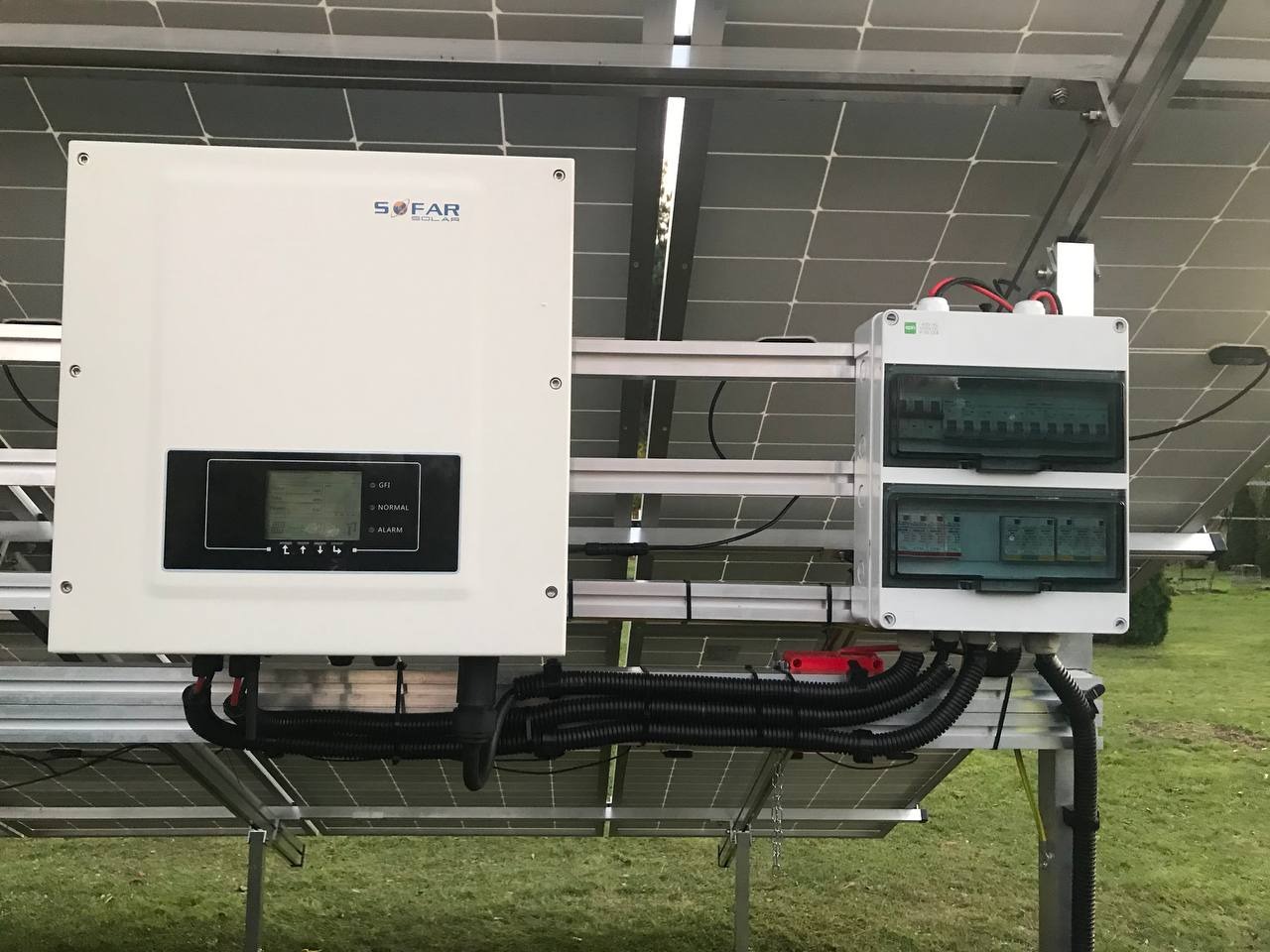Biała skrzynka inwertera SOFAR SOLAR i rozdzielnica elektryczna zamontowane na konstrukcji paneli fotowoltaicznych, widoczne czarne peszle z kablami.