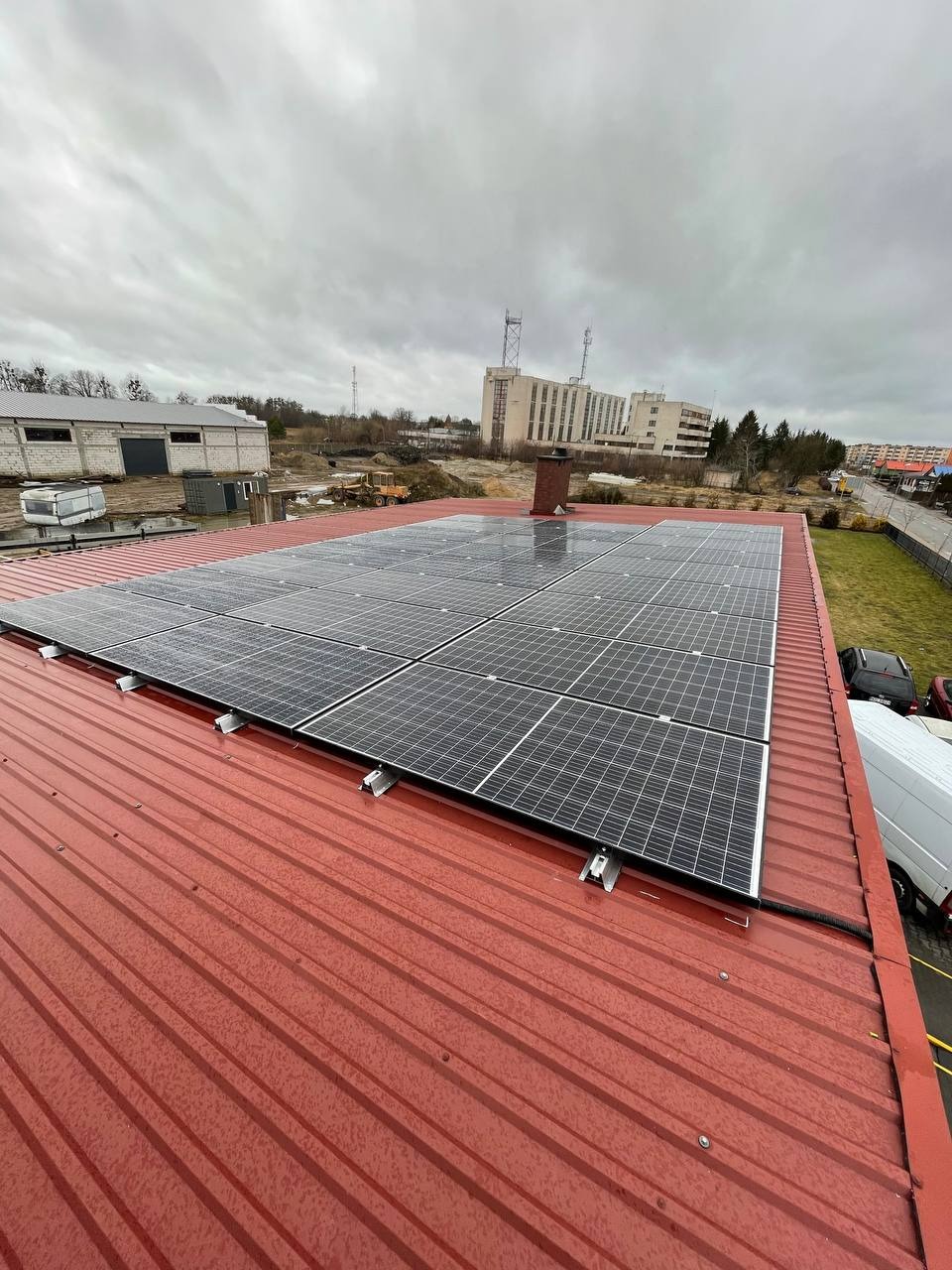 Panele fotowoltaiczne na czerwonym, metalowym dachu budynku przemysłowego w pochmurny dzień, widok z góry.