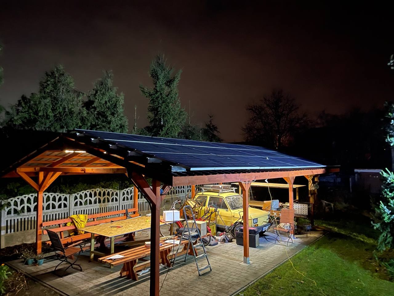 Zadaszenie z panelami fotowoltaicznymi nad tarasem z żółtym samochodem w tle, oświetlone nocą, widok z zewnątrz.