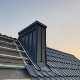 Częściowo pokryty dach z czarną dachówką i kominem wentylacyjnym, widoczna konstrukcja drewniana i membrana w tle niebo o zachodzie słońca.