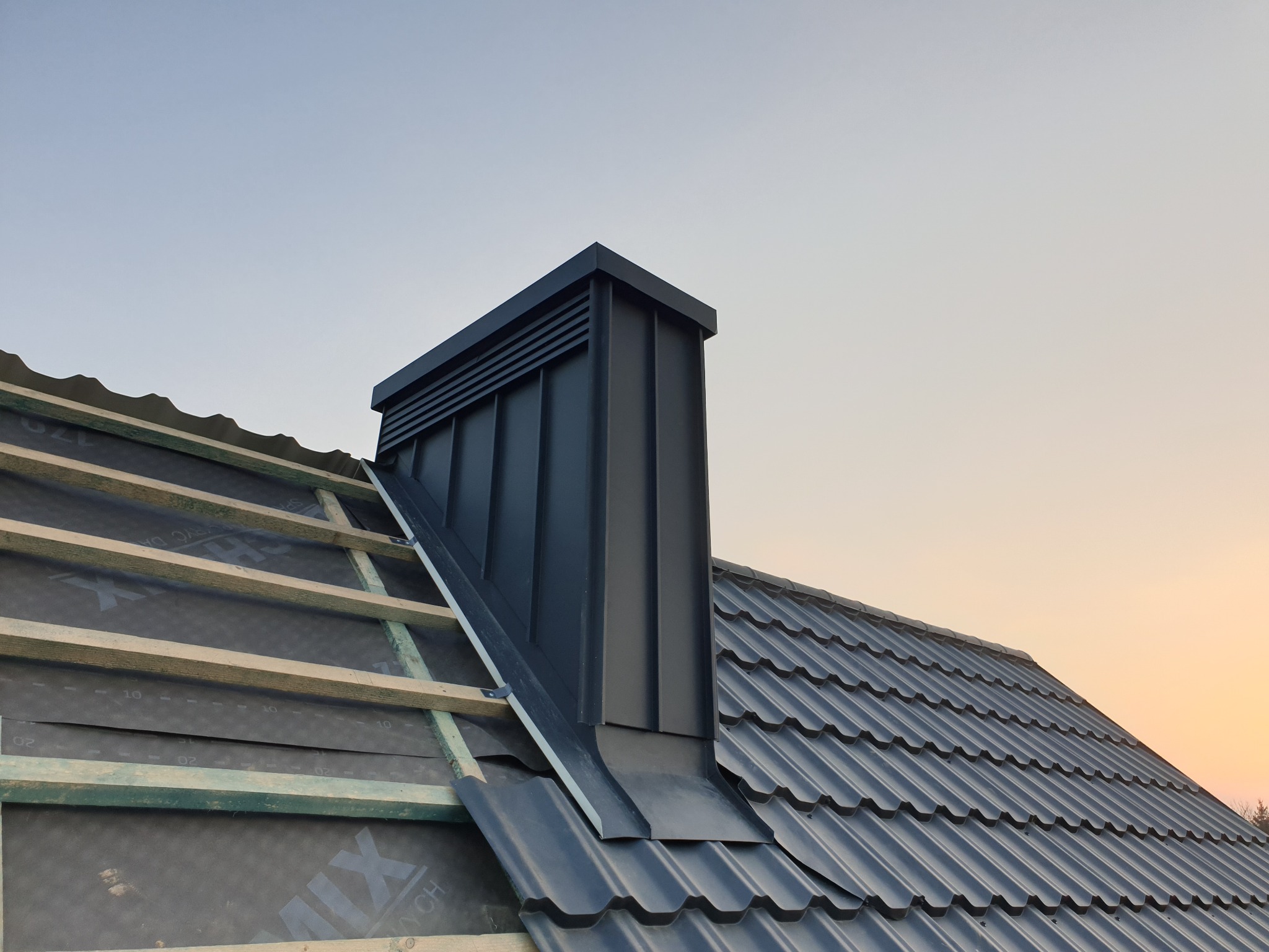 Częściowo pokryty dach z czarną dachówką i kominem wentylacyjnym, widoczna konstrukcja drewniana i membrana w tle niebo o zachodzie słońca.