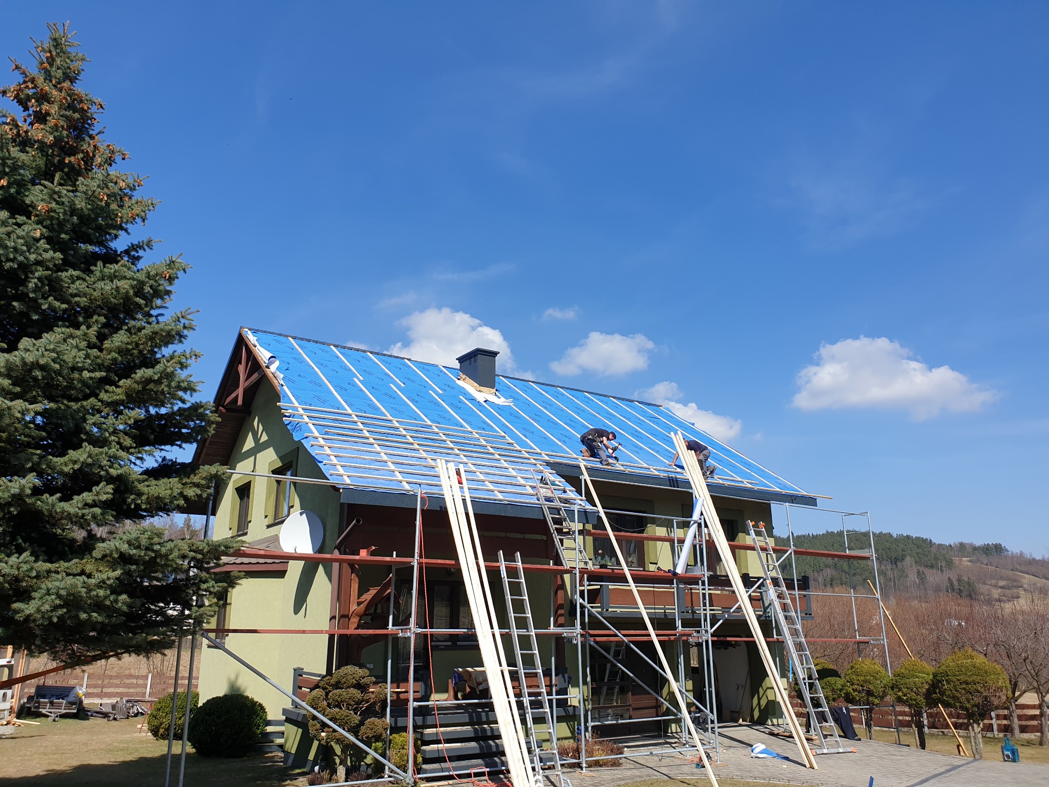 Dwóch dekarzy na rusztowaniu montuje łatowanie na pokrytym niebieską membraną dachu domu jednorodzinnego w słoneczny dzień.