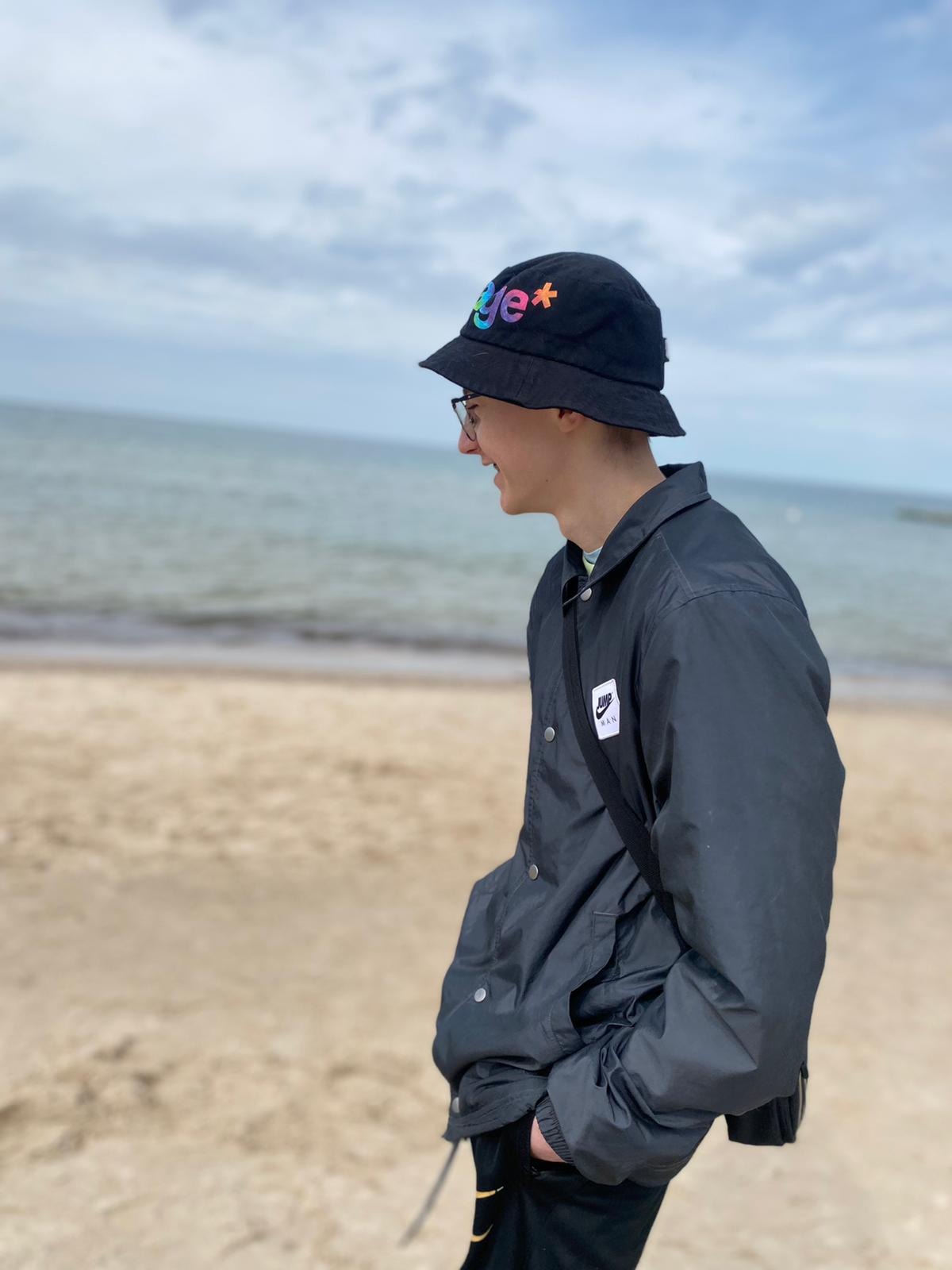 Młody mężczyzna w czarnej kurtce i kapeluszu z kolorowym napisem, spaceruje po piaszczystej plaży z widokiem na morze w pochmurny dzień.