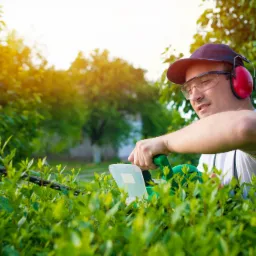 Mężczyzna w okularach ochronnych i słuchawkach przycina żywopłot elektrycznymi nożycami, w tle zielona roślinność i promienie słońca.