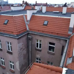 Widok z góry na budynek z czerwonym dachem z dachówki karpiówki, czarną papą wokół kominów i oknem dachowym, elewacja budynku częściowo zniszczona, w tle zabudowa miejska.