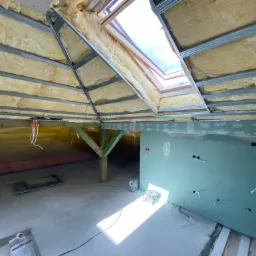 Poddasze w trakcie ocieplania wełną mineralną, widoczne metalowe profile i fragment zamontowanej płyty gipsowo-kartonowej z wyprowadzonymi kablami elektrycznymi oraz okno dachowe.