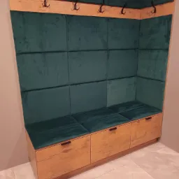 Wnęka z siedziskiem i wieszakami w przedpokoju, obita zielonym welurem, z trzema drewnianymi szufladami na dole i rzędem haczyków na górze, oświetlona ciepłym światłem.