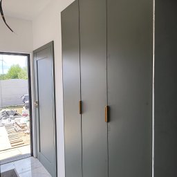 Mk meble Mariusz koczynasz - Szara szafa wnękowa z złotymi uchwytami, widok z boku. W tle widoczne drzwi i plac budowy z paletami, co sugeruje świeżo wykończone wnętrze.