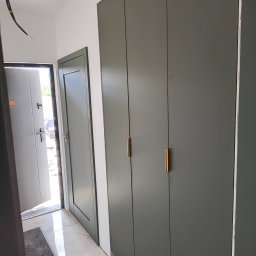 Mk meble Mariusz koczynasz - Nowoczesna, szara szafa wnękowa z złotymi uchwytami, widoczna w korytarzu z otwartymi drzwiami wejściowymi i wewnętrznymi. Minimalistyczny design, płytki podłogowe imitujące marmur.