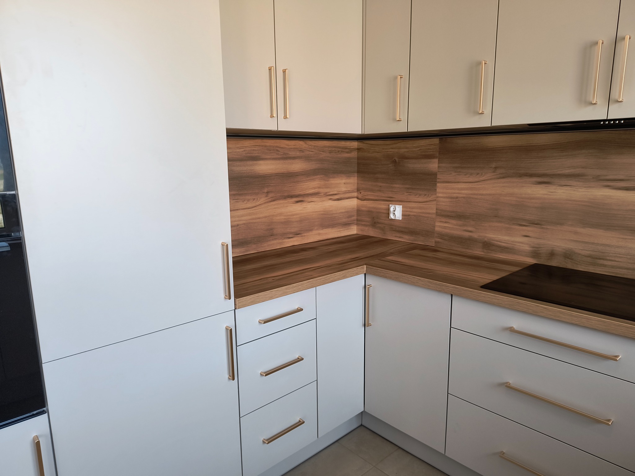 Jasna kuchnia z drewnianym blatem i złotymi uchwytami. Widoczna płyta indukcyjna i gniazdko elektryczne. Górne szafki w odcieniu beżu.