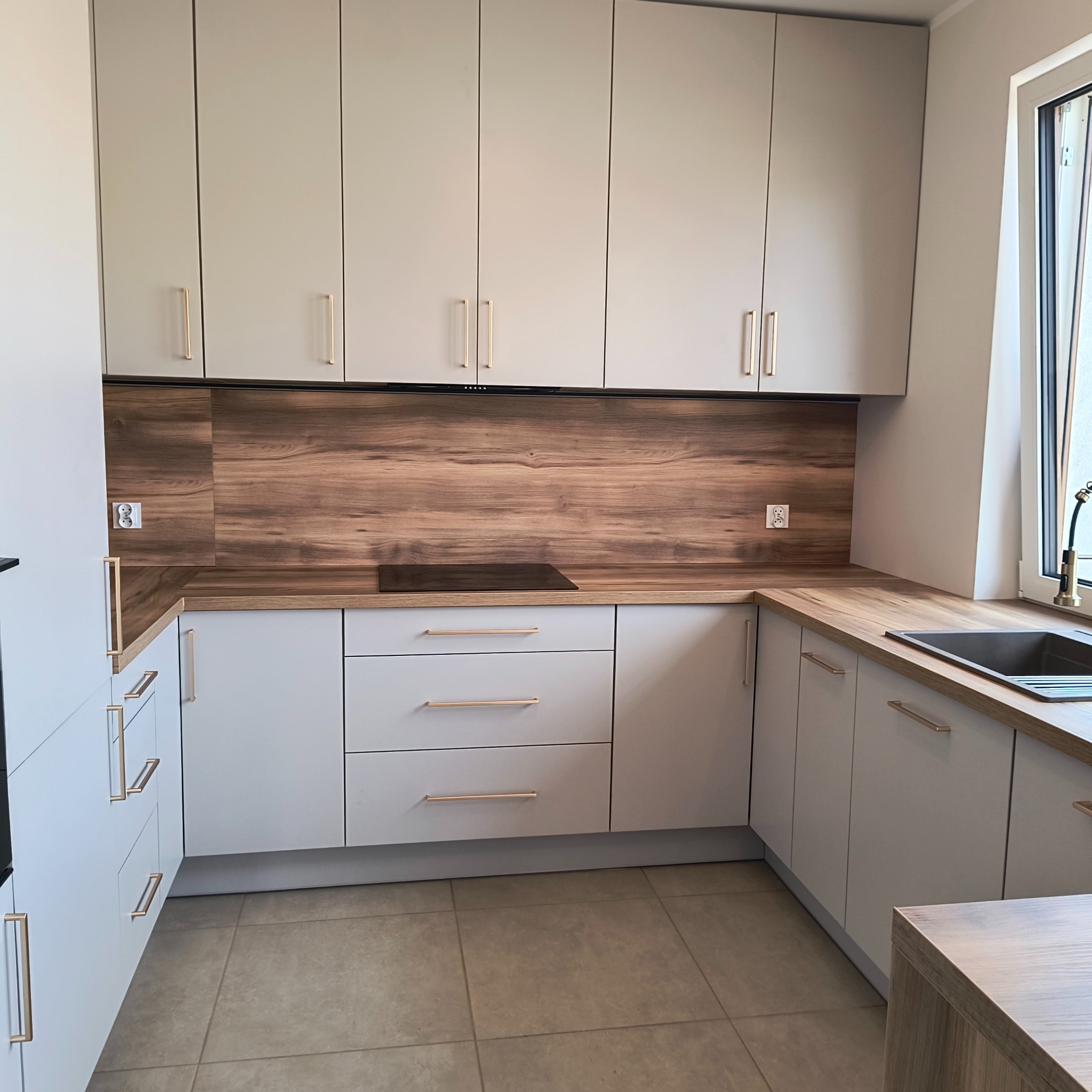 Jasna kuchnia z drewnianym blatem i panelami. Proste, złote uchwyty. Nowoczesne wykończenie, widok na okno i płytę indukcyjną. Minimalistyczny design.