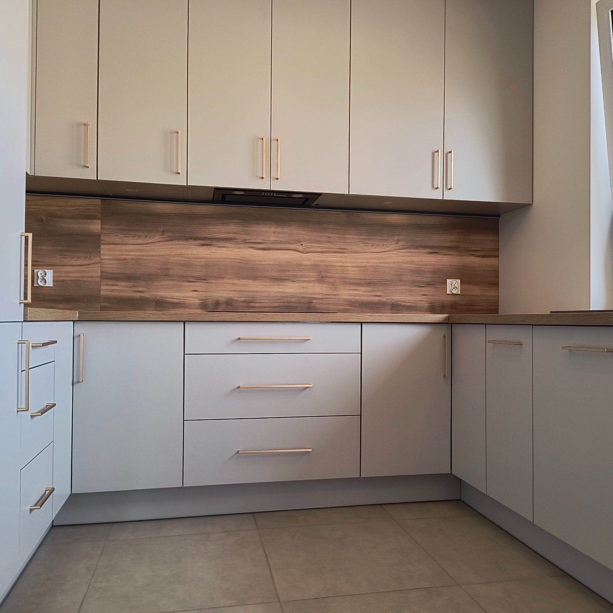 Jasnoszara kuchnia z drewnianym panelem nad blatem i złotymi uchwytami. Widoczne szafki górne i dolne oraz płyta indukcyjna. Nowoczesny design.