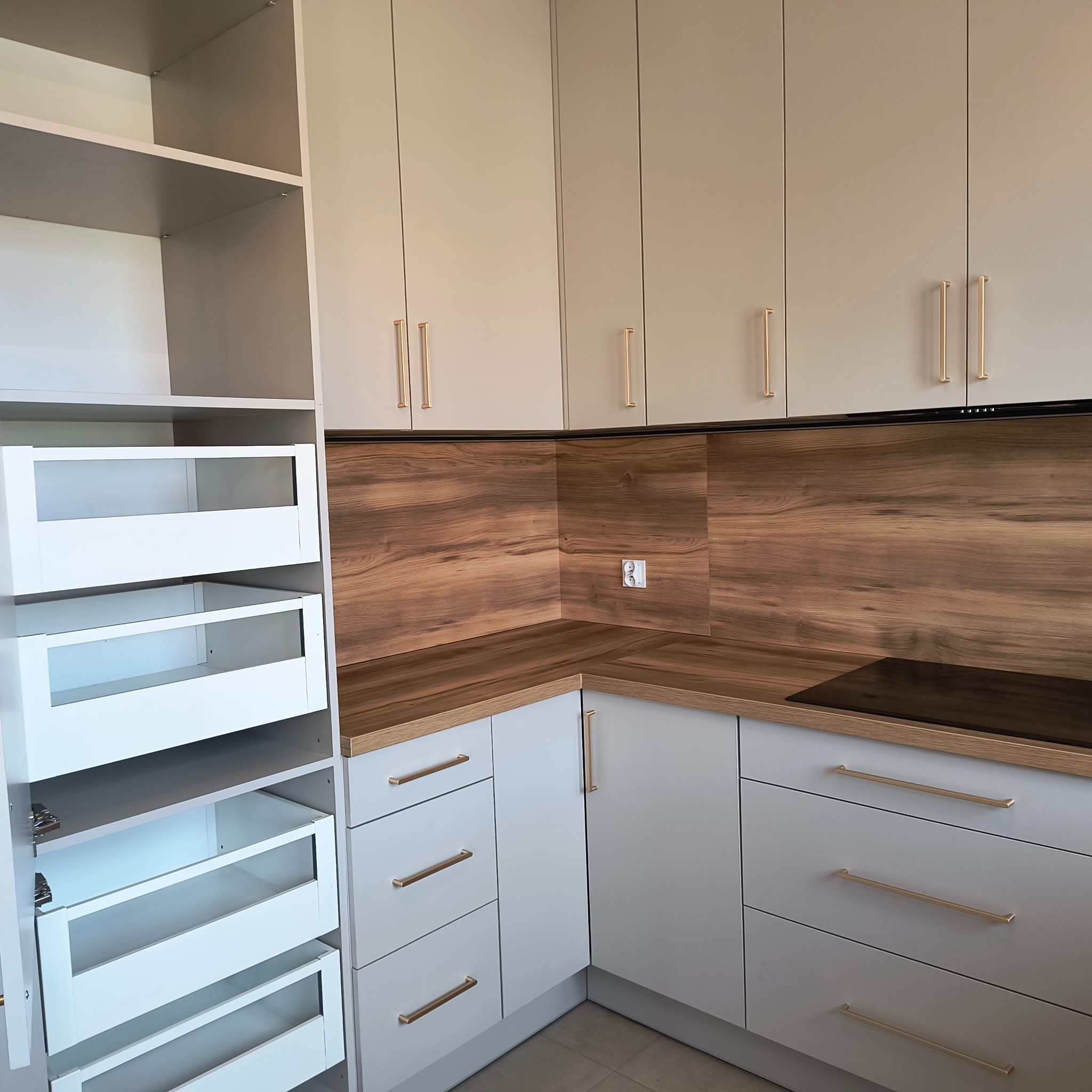 Jasnoszara kuchnia narożna z drewnianym blatem i złotymi uchwytami. Wysoka szafka cargo z wysuwanymi koszami. Nowoczesny design.
