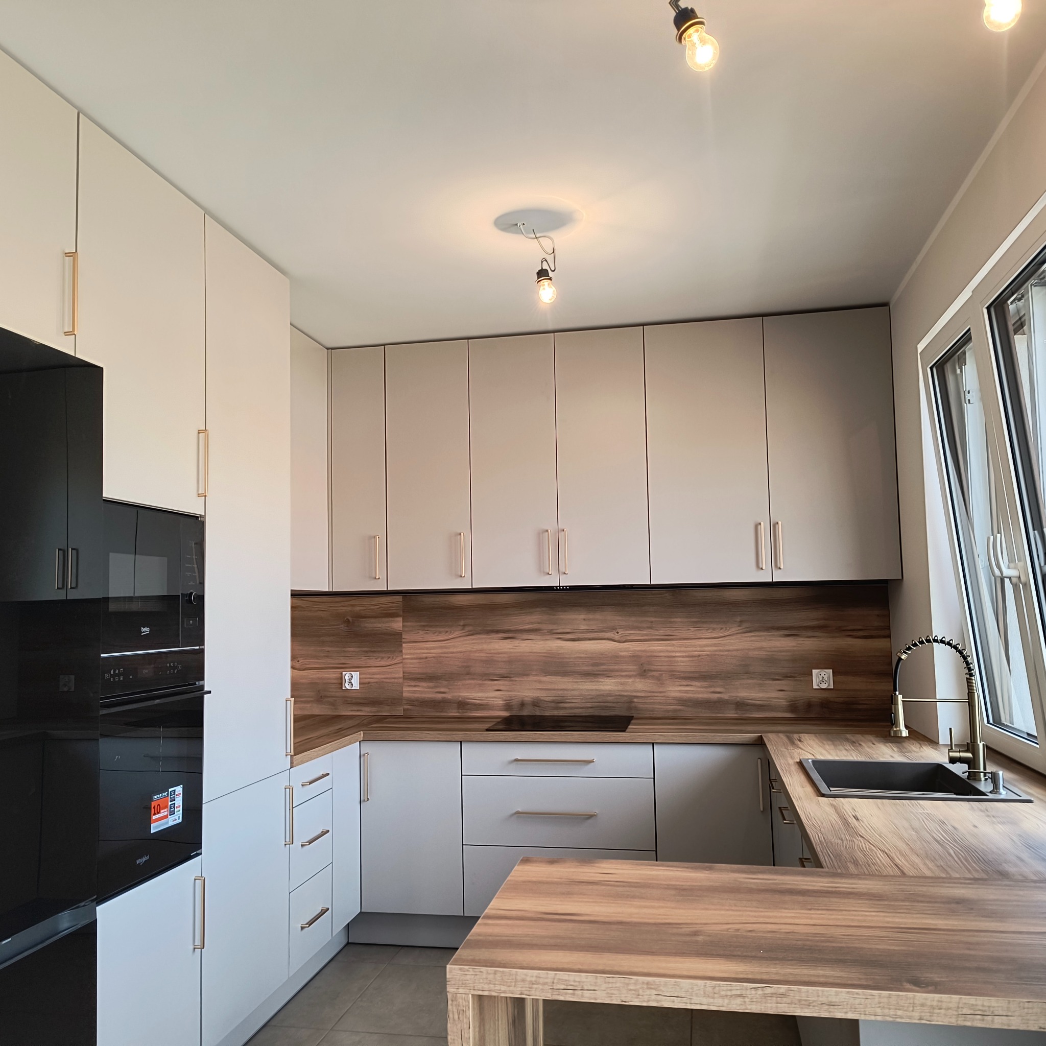 Nowoczesna kuchnia z białymi szafkami i drewnianym blatem, oświetlona żarówkami. Uwagę przykuwa czarny piekarnik i płyta grzewcza. Widoczne okno wpuszcza naturalne światło.