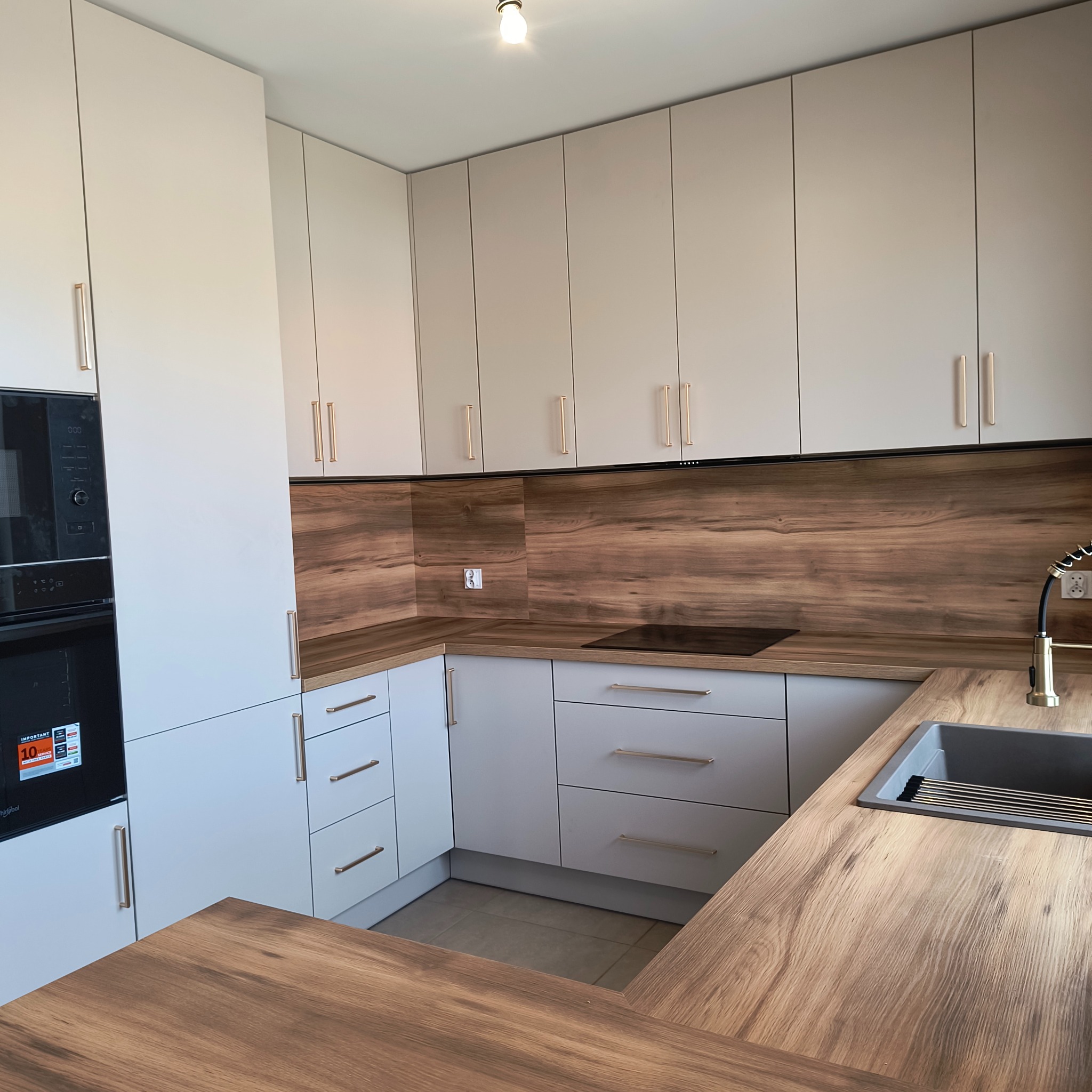 Jasna kuchnia z drewnianym blatem i złotymi uchwytami. Widoczny zlew, płyta indukcyjna i zabudowana kuchenka mikrofalowa.