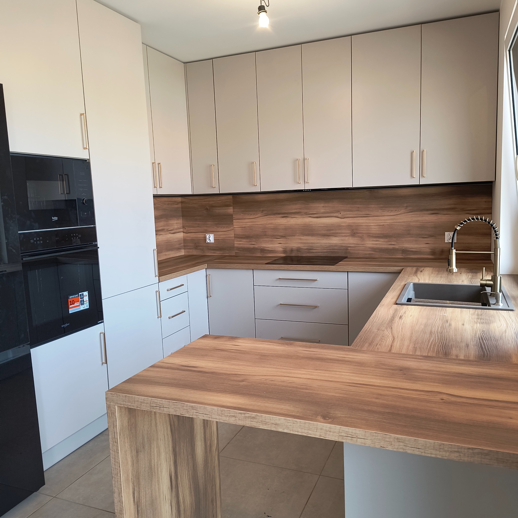 Jasna kuchnia z drewnianym blatem i panelami, fronty szafek w odcieniach bieli i szarości, złote uchwyty, nowoczesny design, oświetlenie punktowe.