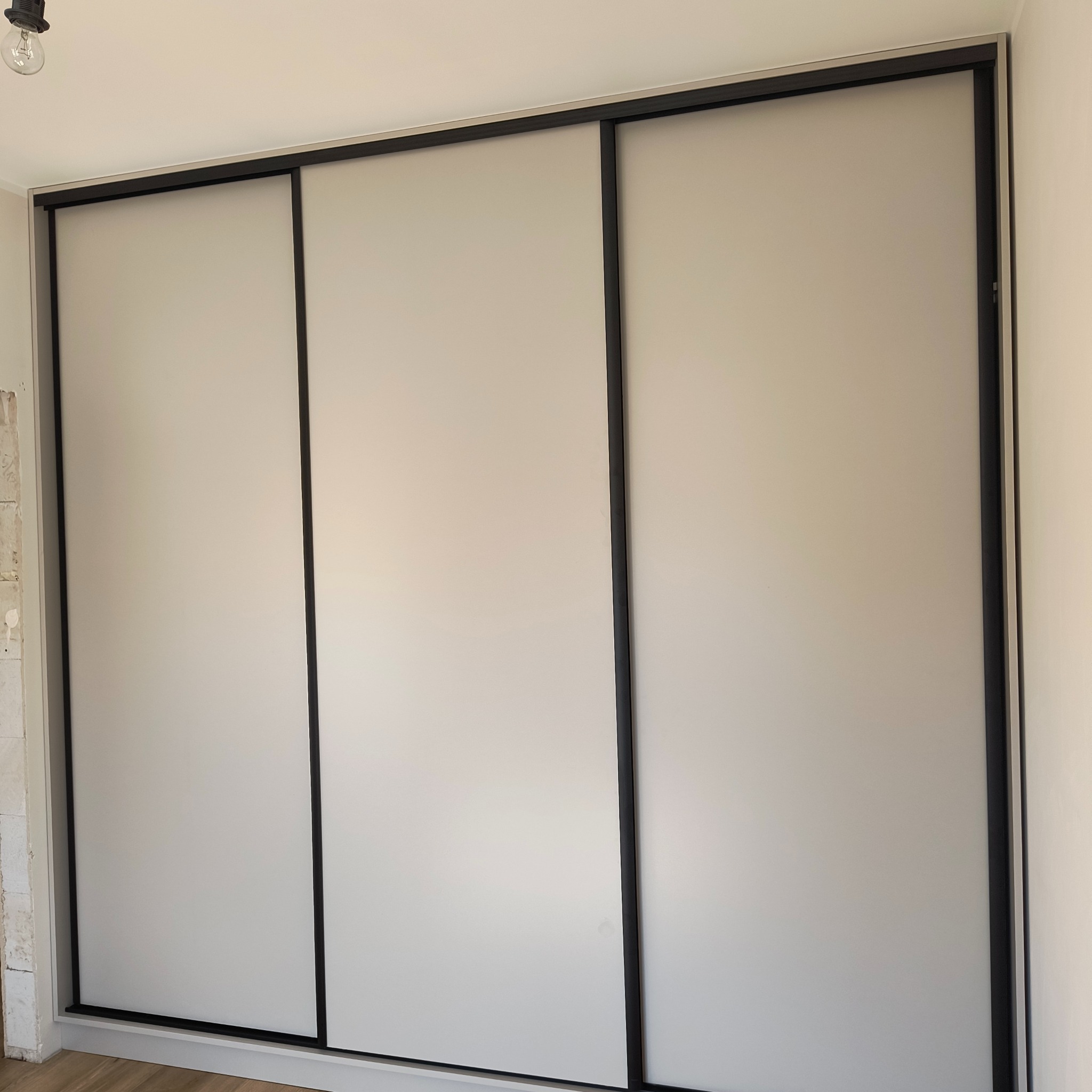 Minimalistyczna szafa wnękowa z przesuwnymi, białymi drzwiami i czarnymi ramami, widziana z bliska, z surowym wykończeniem ściany po lewej stronie.