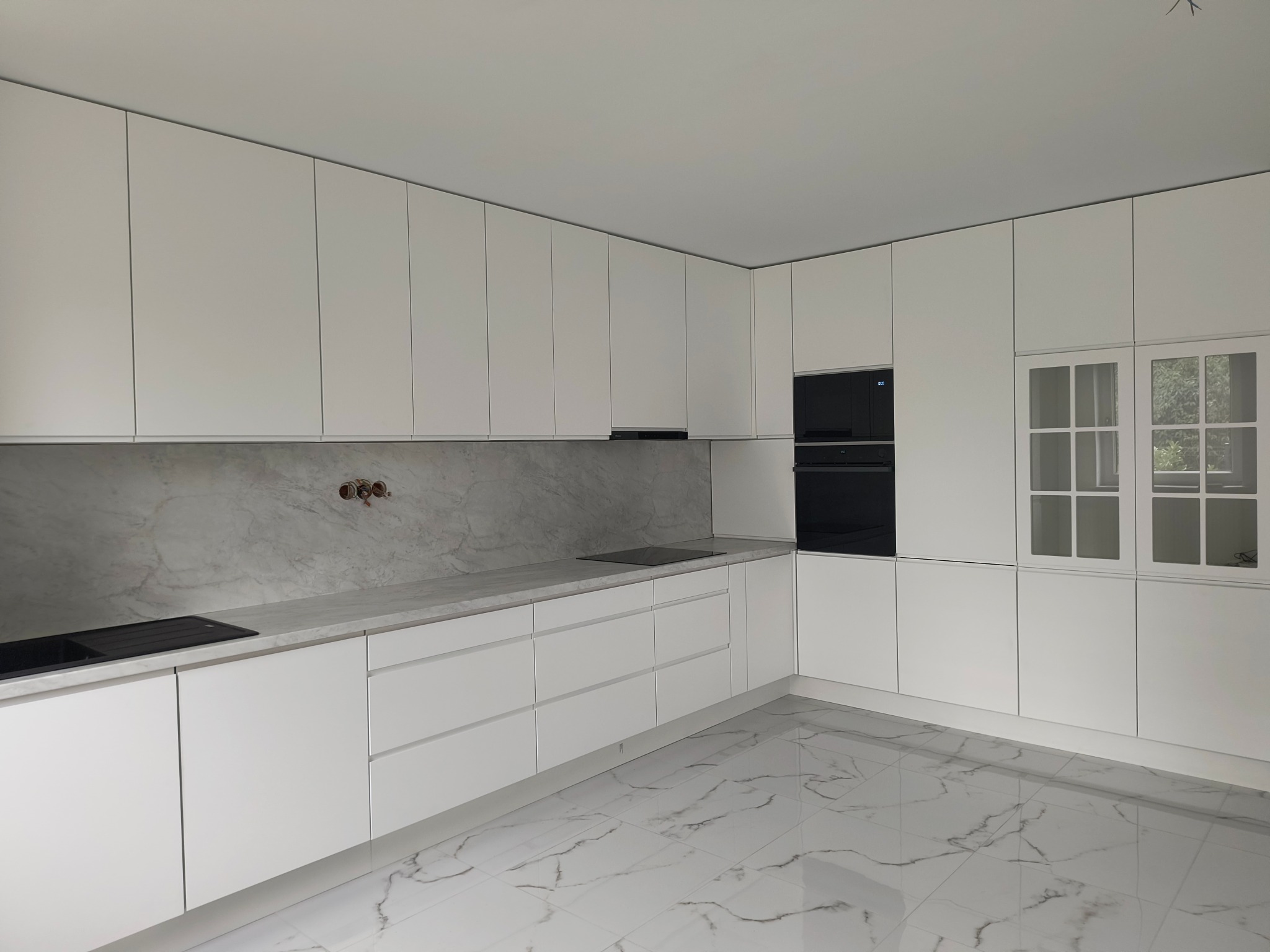 Nowoczesna, minimalistyczna kuchnia z białymi, gładkimi frontami i marmurowym blatem. W tle okno z białymi szprosami i sprzęt AGD w zabudowie. Podłoga z białych płytek imitujących marmur.