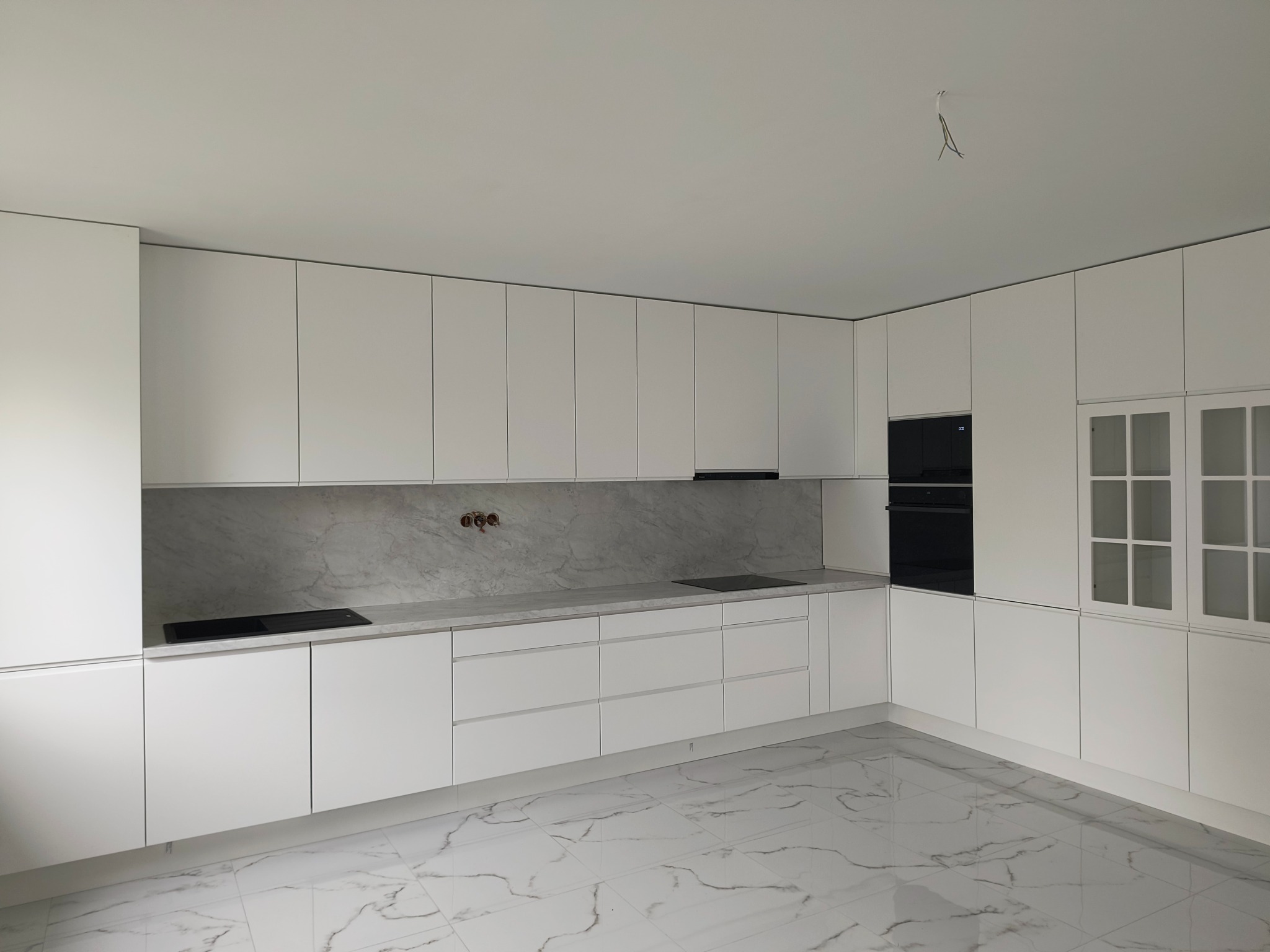 Minimalistyczna kuchnia w Miłowicach z białymi frontami i marmurowym blatem. Widoczne płyty grzewcze i szklane szafki, całość w jasnej tonacji.