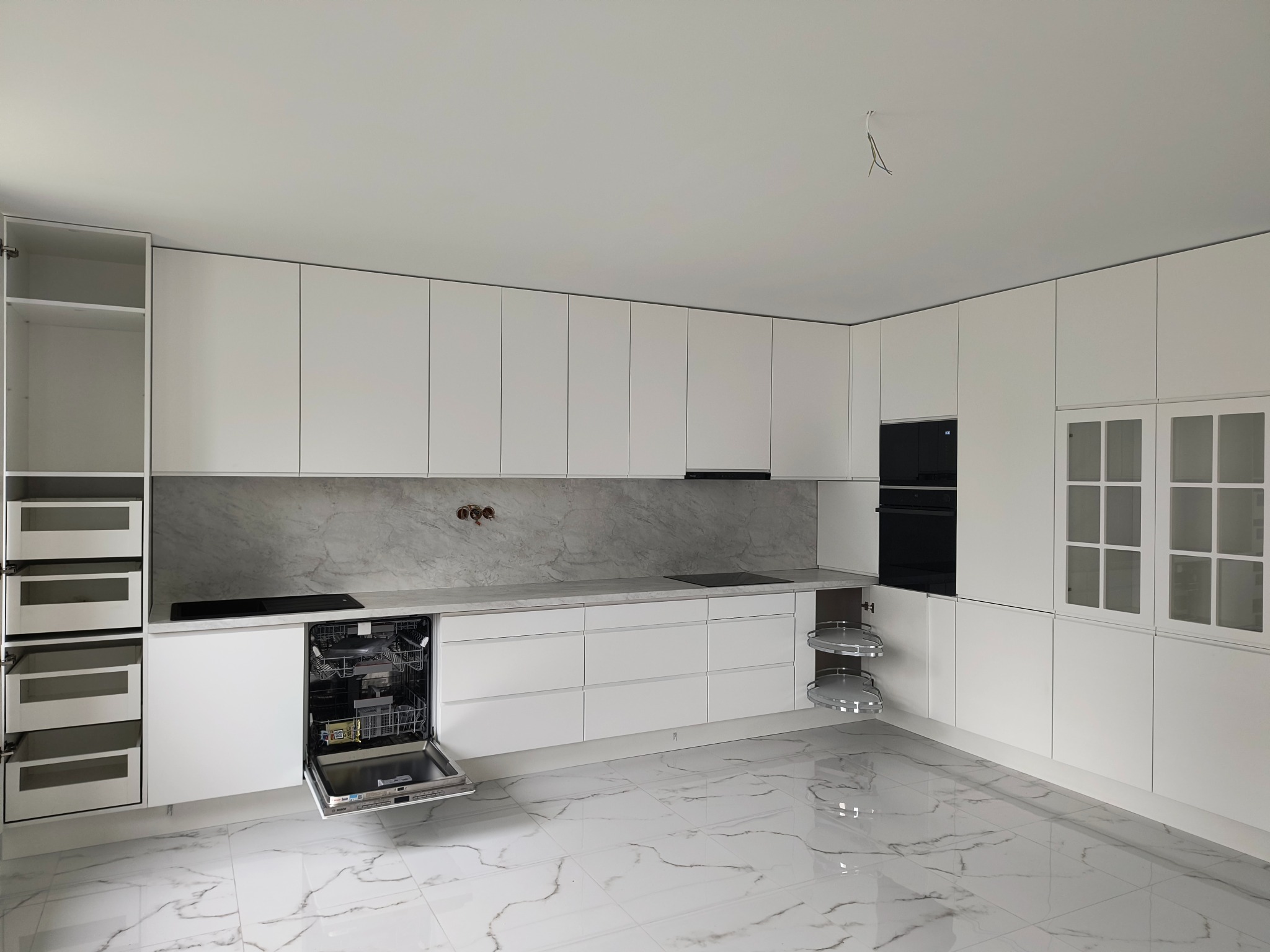 Nowoczesna, jasna kuchnia z białymi szafkami i marmurowym blatem. Otwarta zmywarka i wysuwane półki narożne. Podłoga z płytek imitujących marmur.