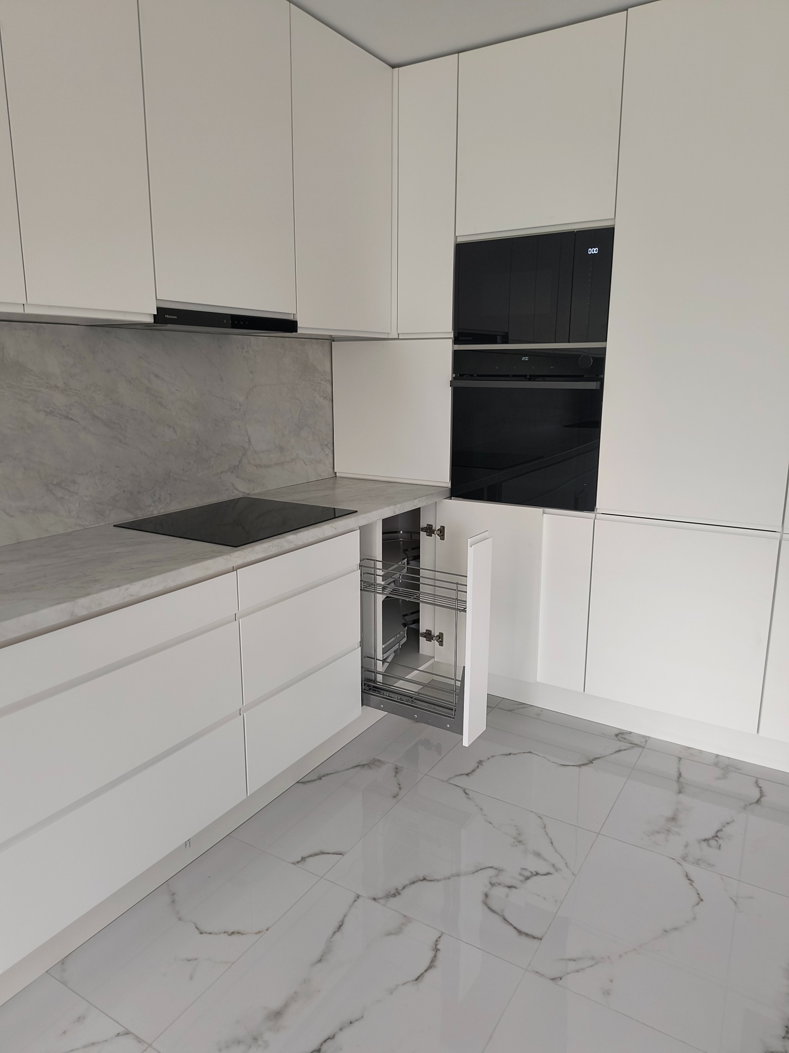 Nowoczesna, minimalistyczna kuchnia w bieli z marmurowym blatem i wysuwanym cargo. Widoczny piekarnik i płyta indukcyjna. Podłoga wyłożona białym gresem.