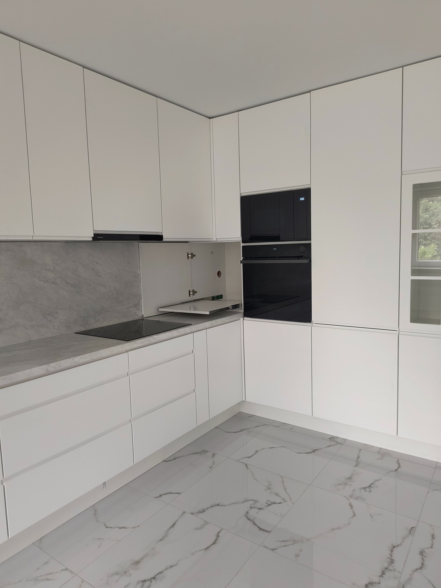 Nowoczesna, minimalistyczna kuchnia z białymi frontami, marmurowym blatem i czarnym sprzętem AGD. Widoczne otwarte szafki i marmurowe płytki na podłodze.