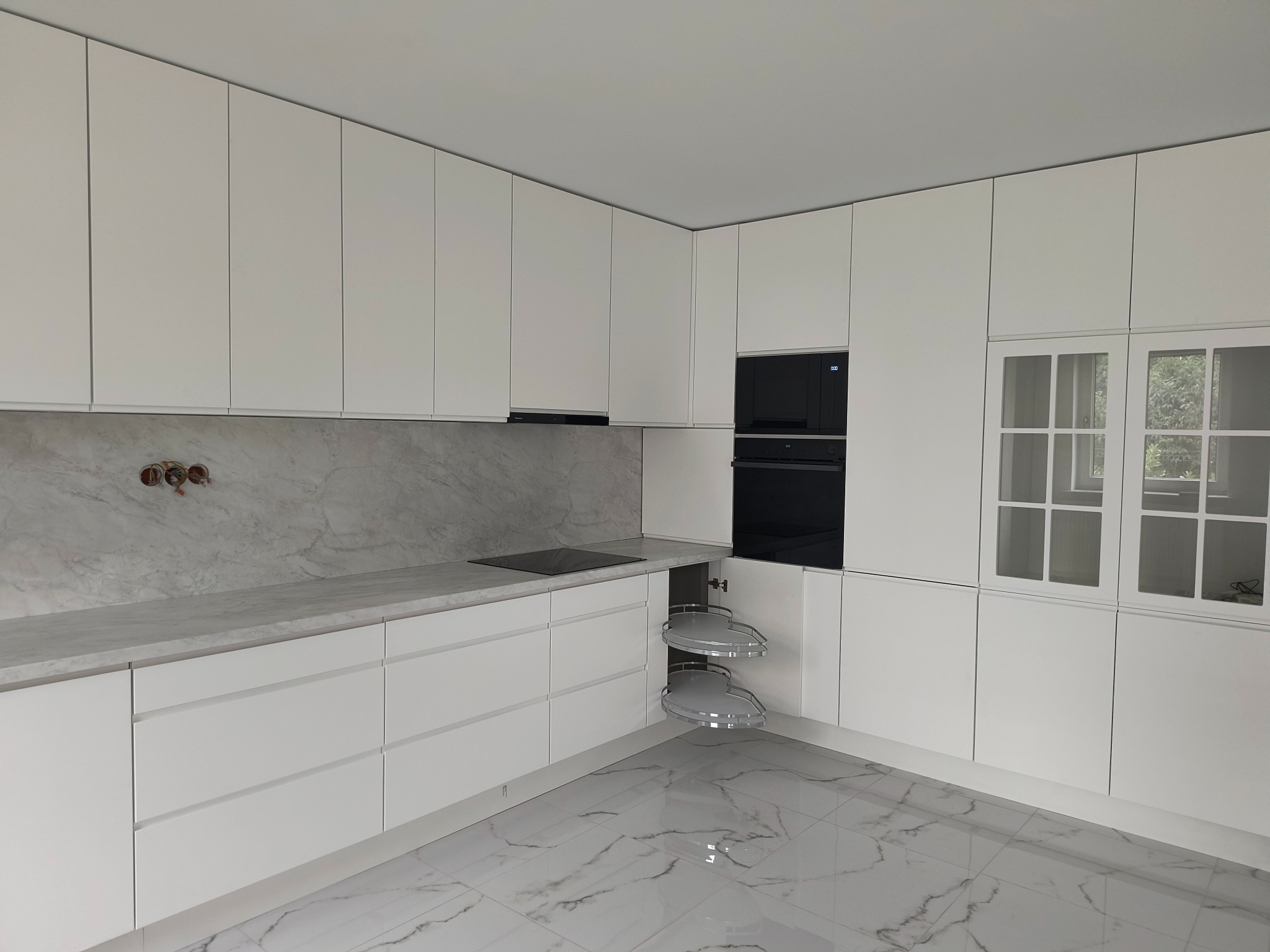 Minimalistyczna, biała kuchnia z marmurowym blatem i podłogą. Uwagę przyciąga szafka narożna z obrotowymi półkami oraz przeszklona witryna w górnej zabudowie.