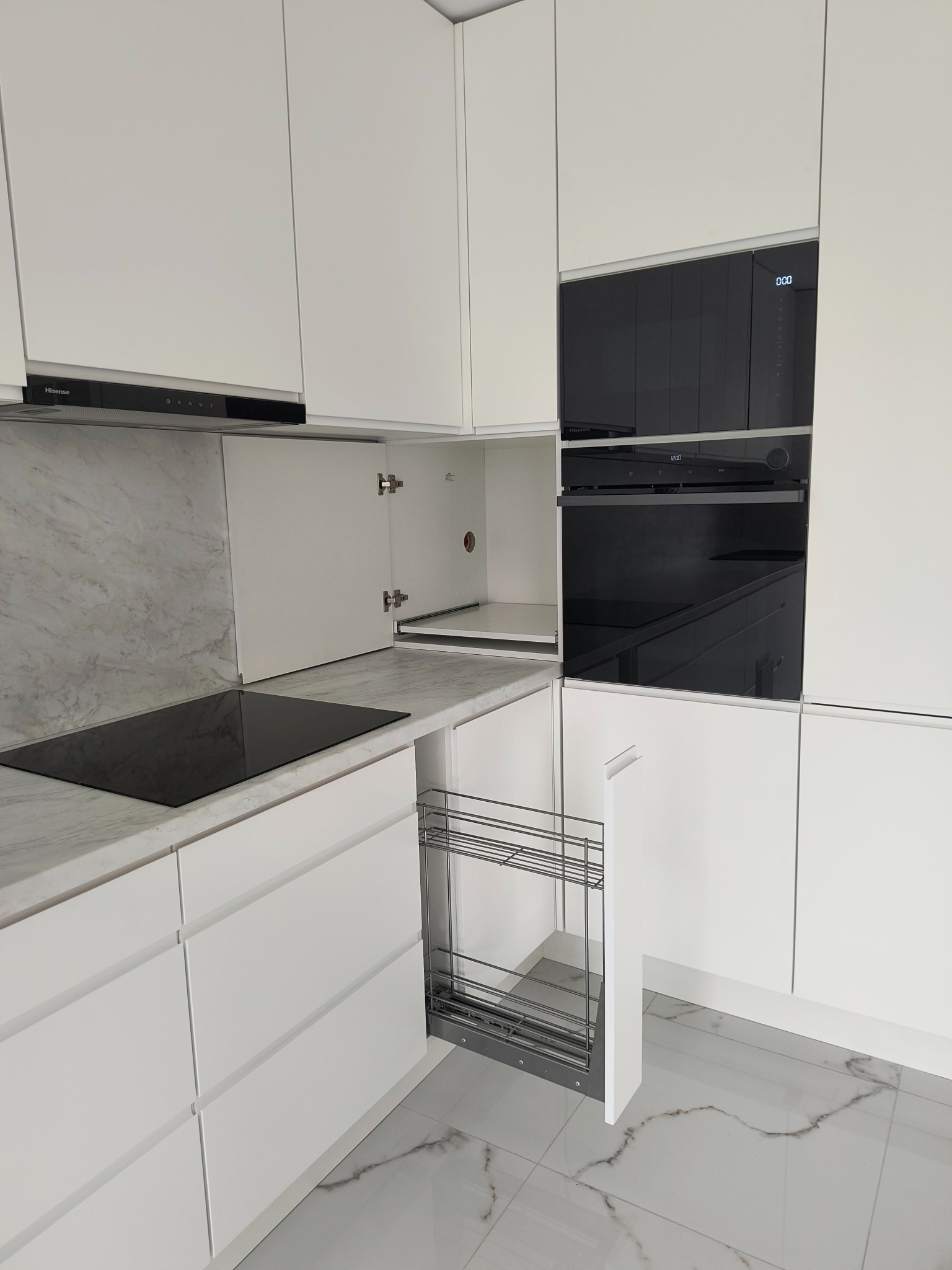 Biała kuchnia na wymiar z otwartą szafką narożną, wysuwanym koszem cargo i czarnym sprzętem AGD. Marmurowy blat i ściana nad blatem.