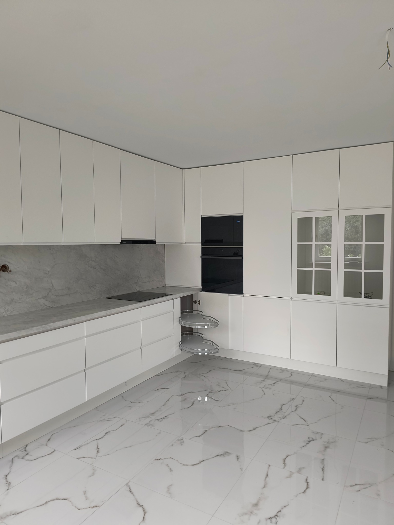 Nowoczesna, biała kuchnia z marmurowym blatem i podłogą. Widoczne szafki z przeszklonym frontem i wysuwany kosz narożny. Minimalistyczny design i jasne wnętrze.