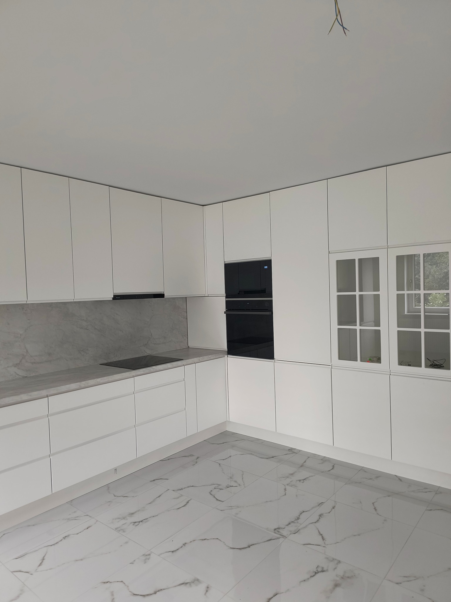 Minimalistyczna, biała kuchnia z marmurowym blatem i podłogą. Zabudowa z piekarnikiem i szafkami z przeszkleniami. Widoczne wystające kable elektryczne.