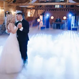 Para młoda tańczy pierwszy taniec w drewnianej sali weselnej, oświetlonej reflektorami i wypełnionej sztuczną mgłą, z DJ-em w tle.