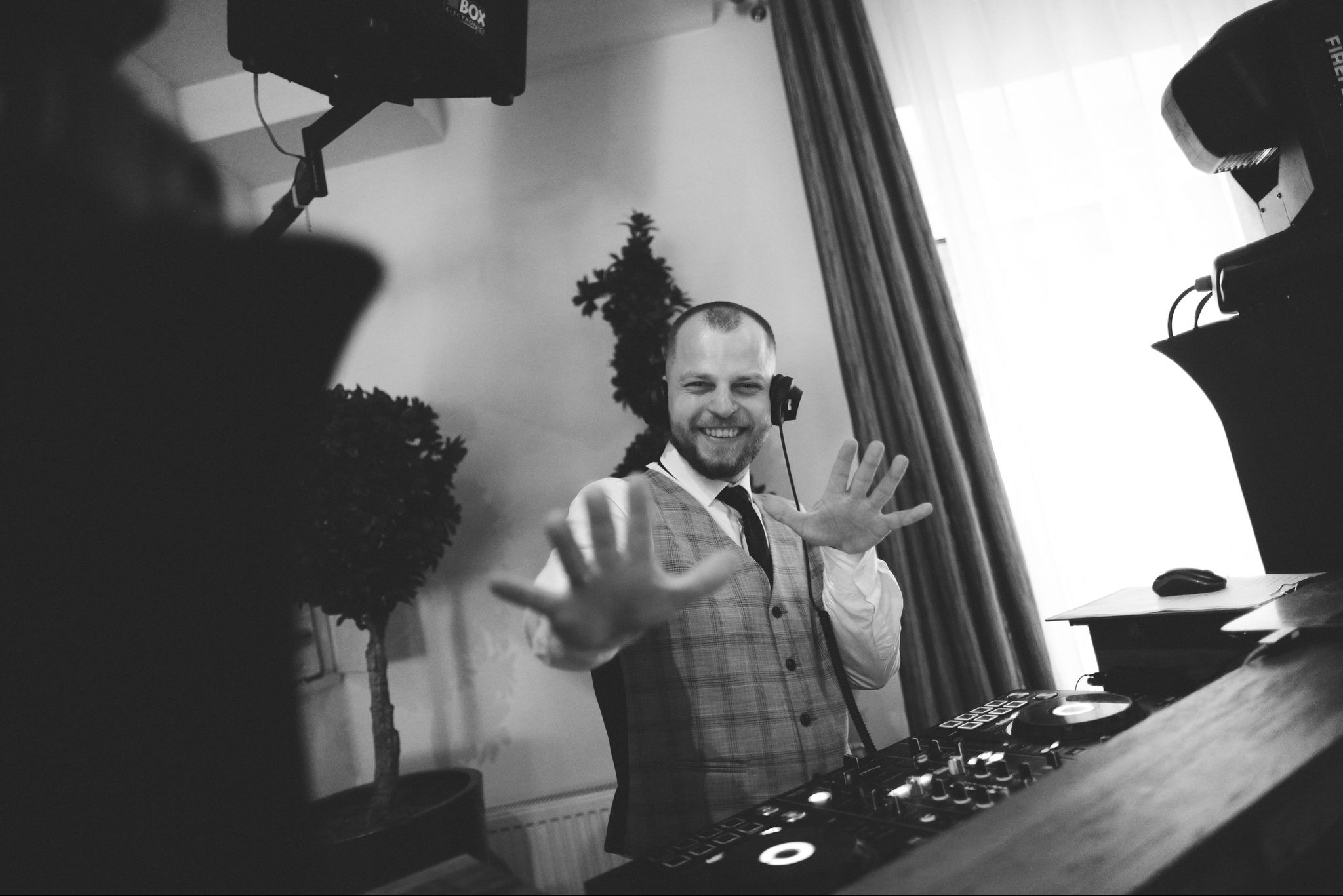 Uśmiechnięty DJ w słuchawkach i kraciastej kamizelce, z rozłożonymi dłońmi za konsoletą, w tle rośliny doniczkowe i oświetlenie sceniczne.