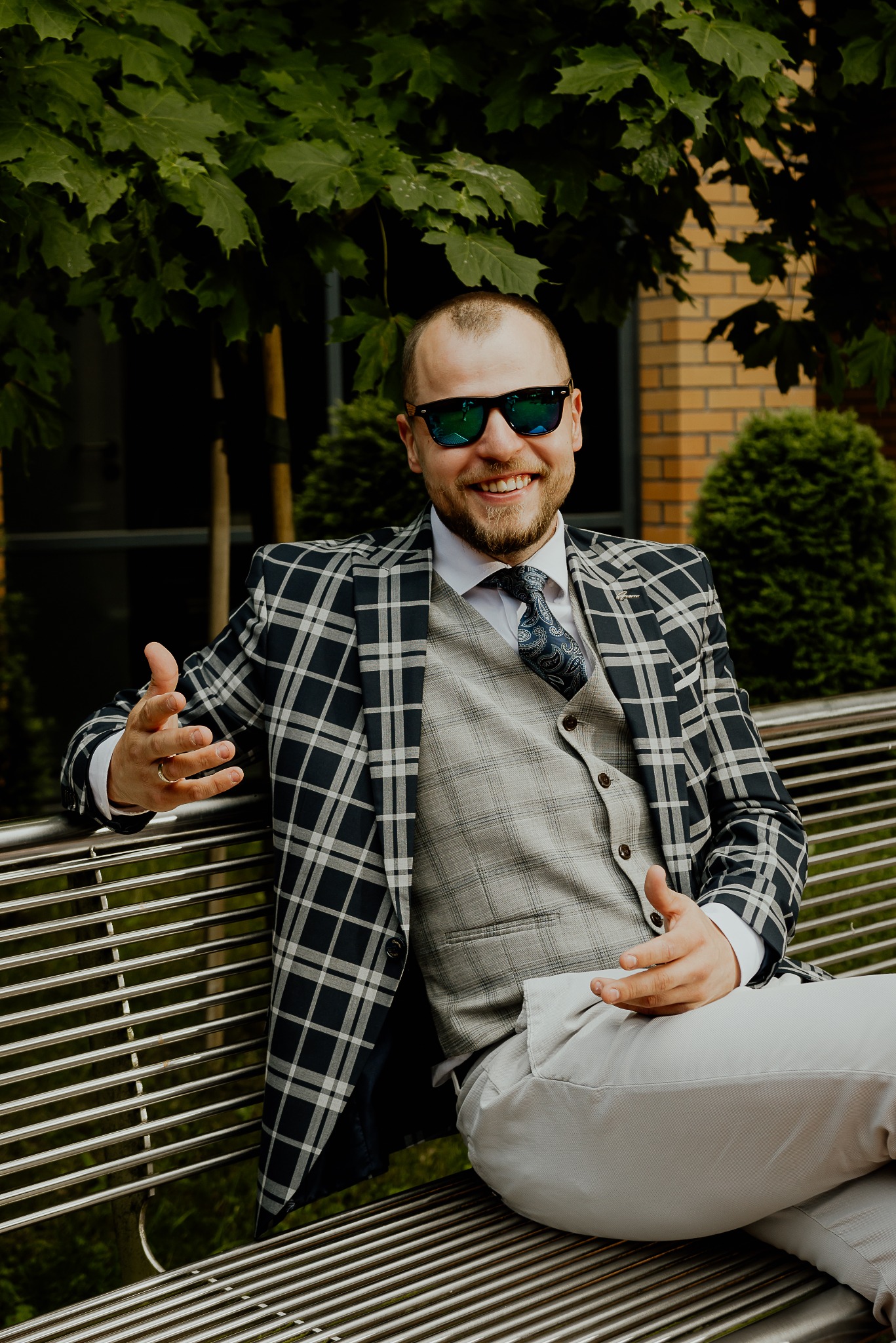 Uśmiechnięty mężczyzna w okularach przeciwsłonecznych, w kraciastej marynarce i kamizelce, siedzi na metalowej ławce na tle zieleni i budynku z cegły.