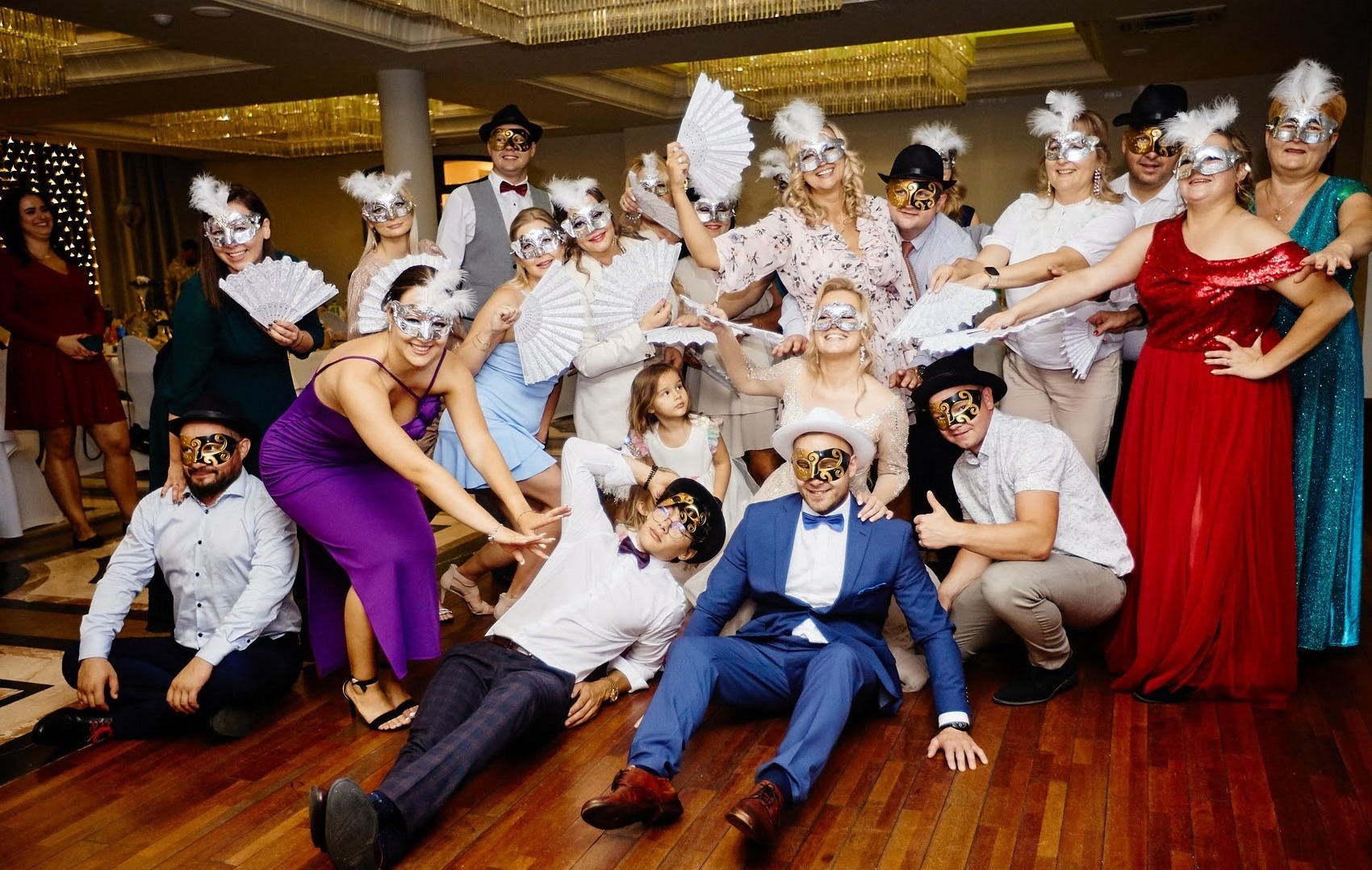 Grupa osób w maskach karnawałowych pozuje do zdjęcia na parkiecie podczas imprezy okolicznościowej, część osób trzyma wachlarze, widoczny żyrandol