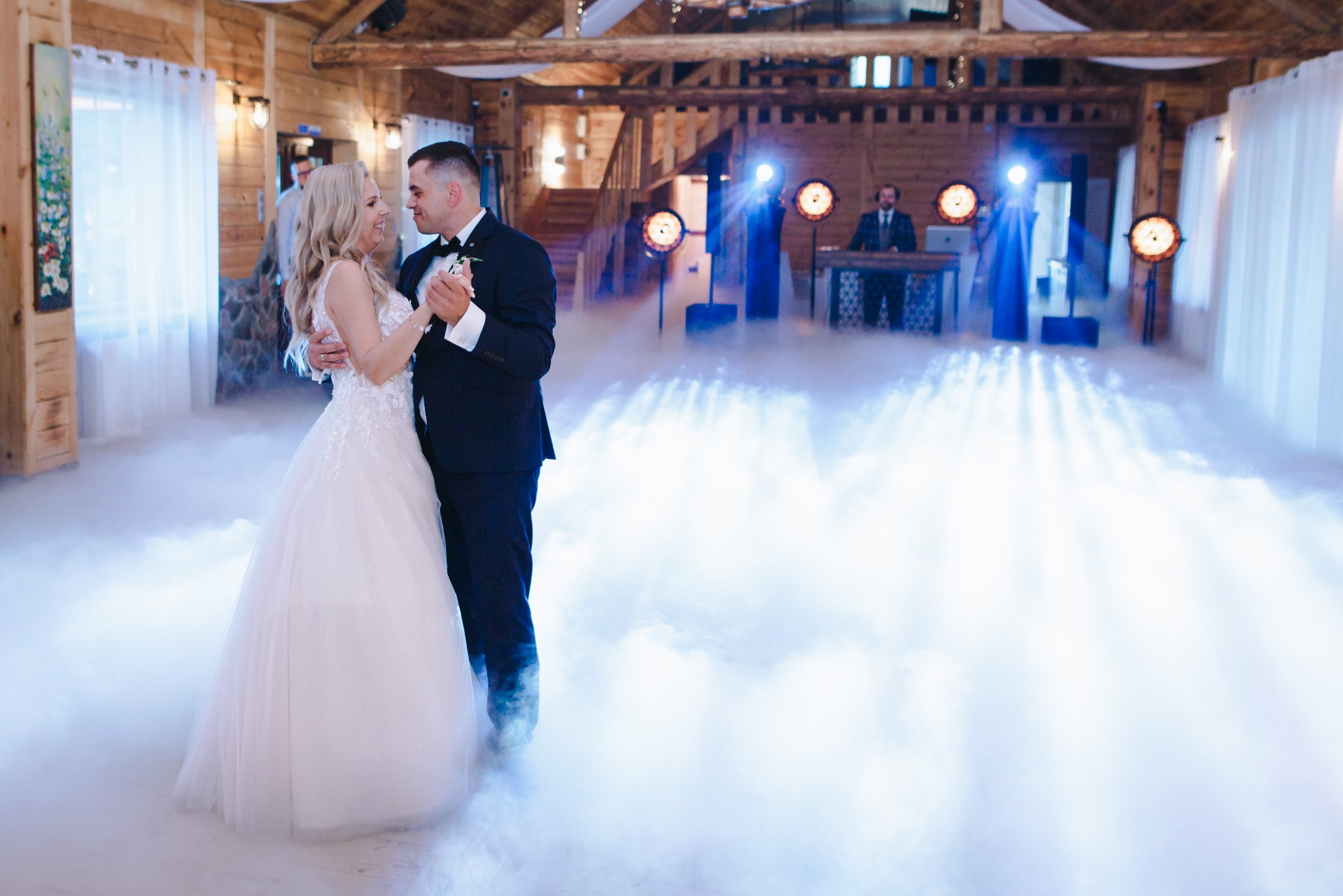 Para młoda tańczy pierwszy taniec w drewnianej sali weselnej, oświetlonej reflektorami i wypełnionej sztuczną mgłą, z DJ-em w tle.