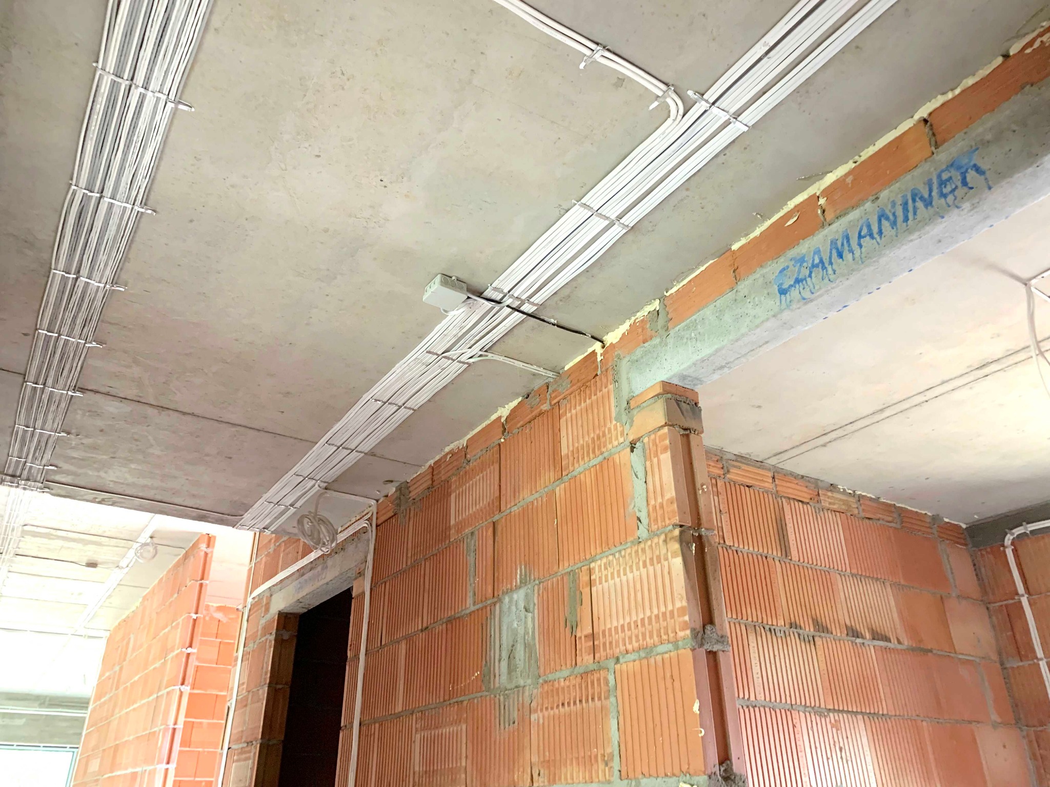 Widok z dołu na sufit z zamocowanymi, białymi przewodami elektrycznymi w rurkach, prowadzonymi wzdłuż betonowego sufitu i ścian z czerwonej cegły dziurawki. Na betonowej belce widoczny napis...